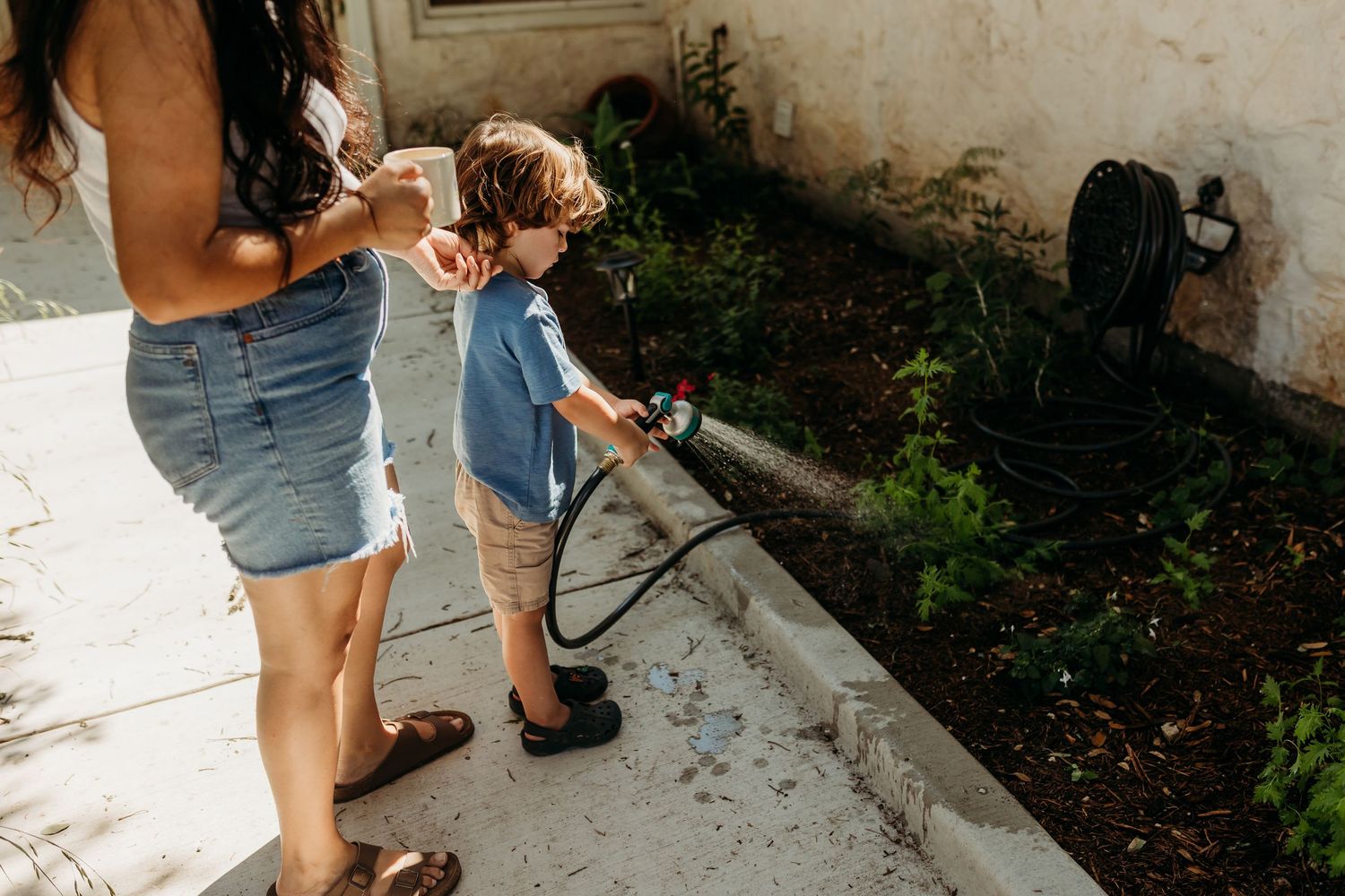 A mother guides her young son as he waters a garden bed with a hose on a sunny day.
