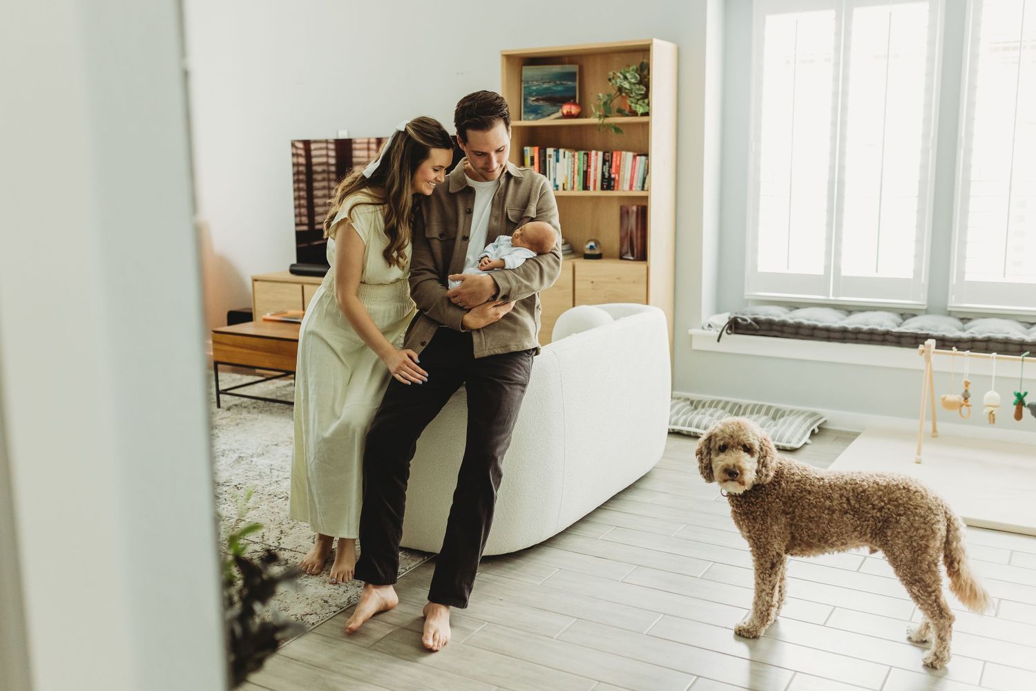 Young couple with newborn baby and fluffy brown dog standing in bright modern living room.