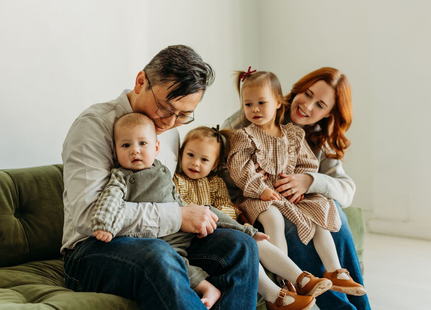 Family of five with three young children sit together on green couch in bright white studio.