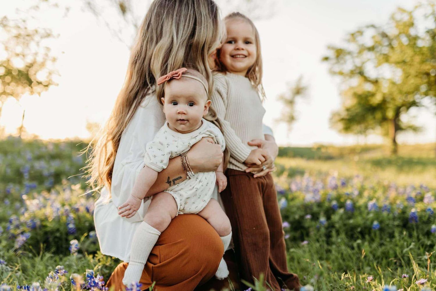 A mother kneels in a bluebonnet field holding her baby while her toddler smiles beside her.