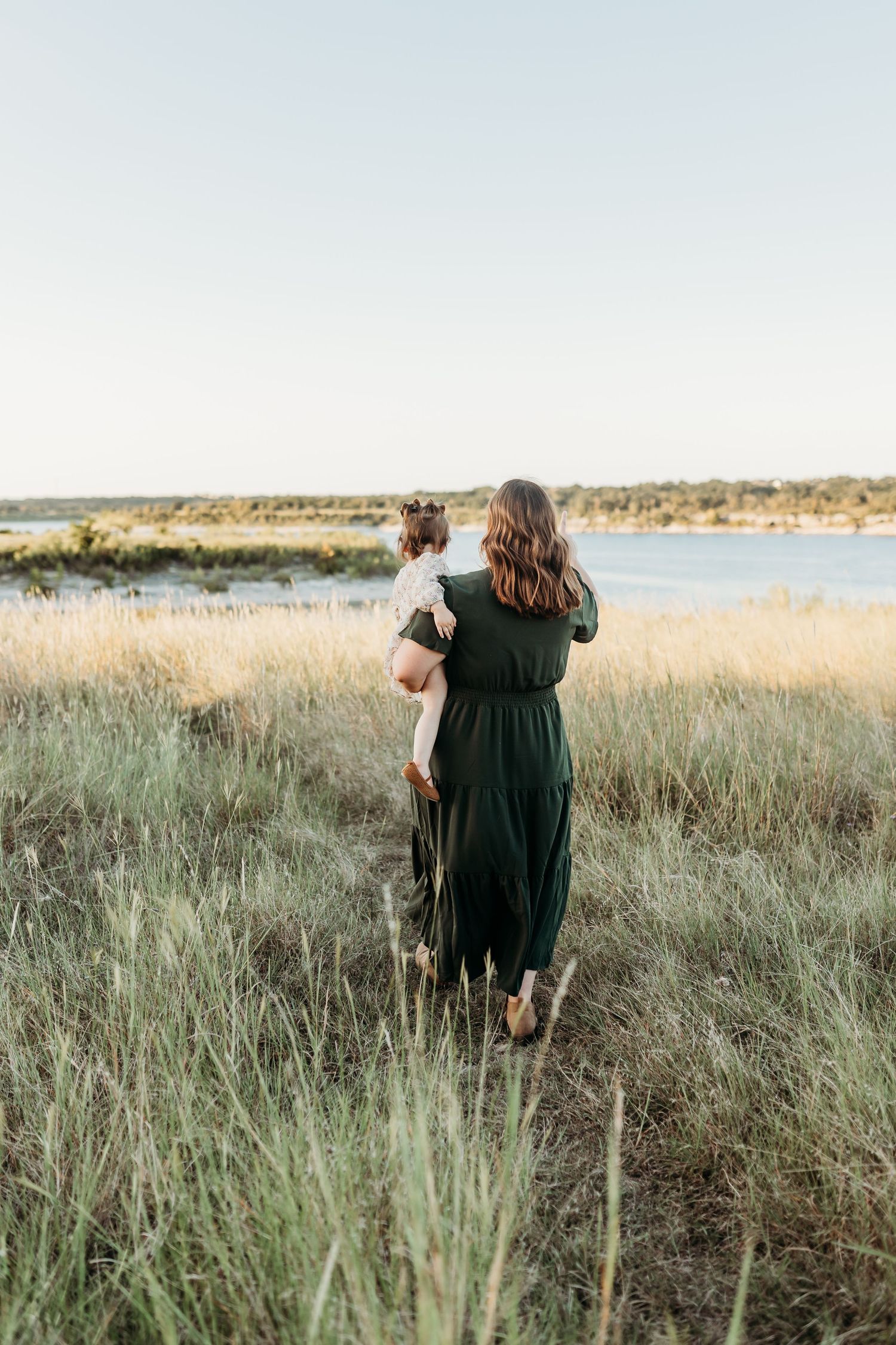 Mother in green jumpsuit holding toddler while standing in tall grass near a lake.
