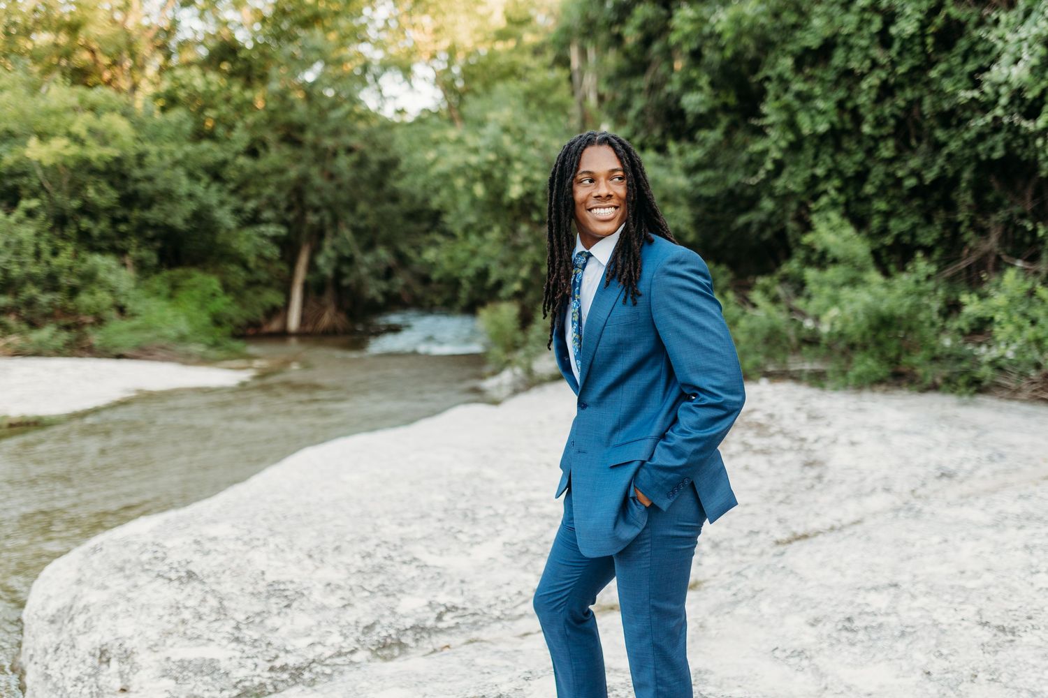 Smiling man in blue suit standing on rocks by a creek outdoors.