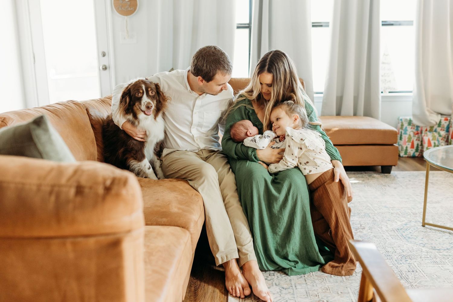 Family with two children and border collie dog sitting together on brown leather couch in bright living room.
