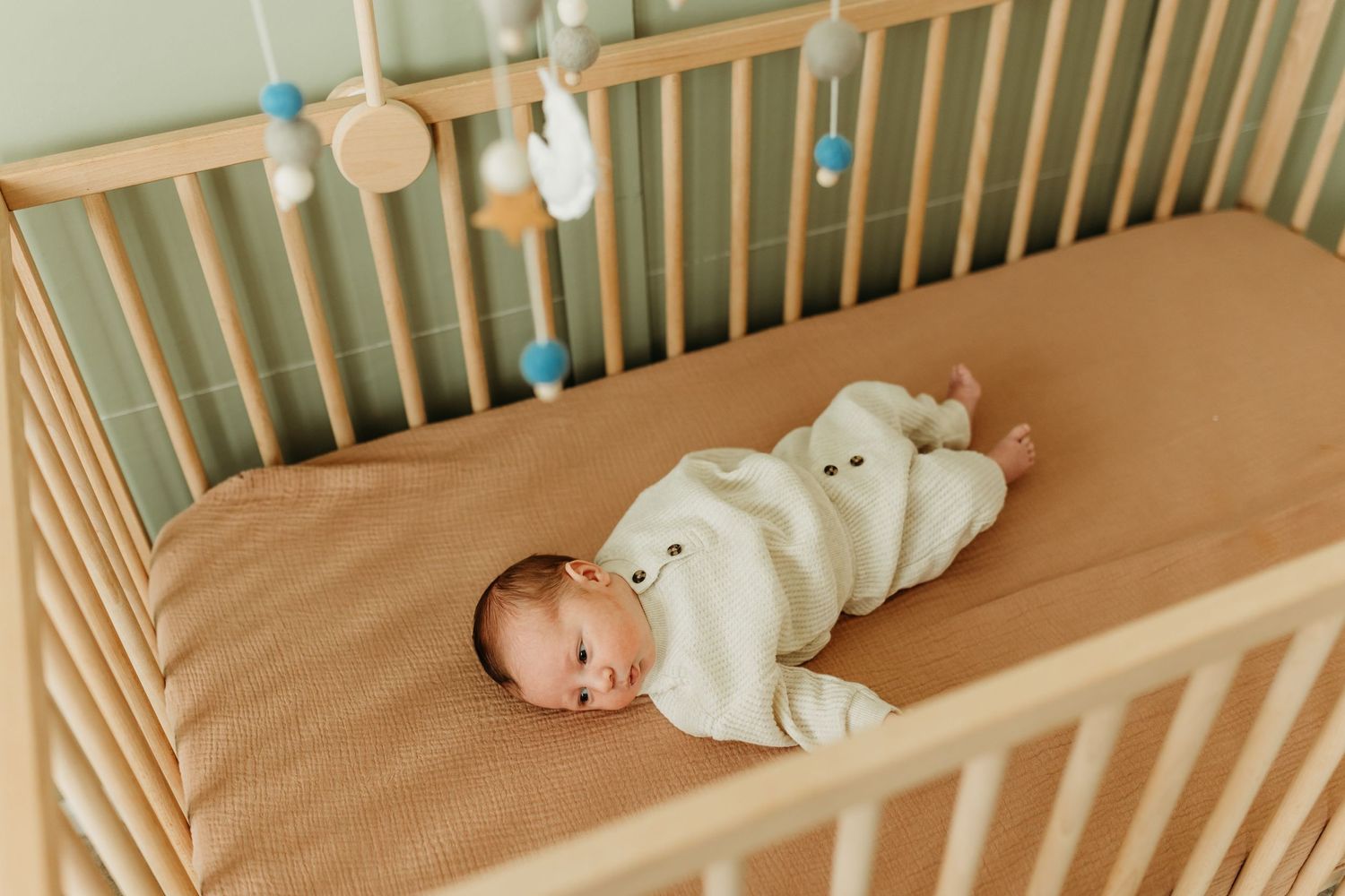 Newborn baby in cream bear outfit lies awake in wooden crib with mobile above.