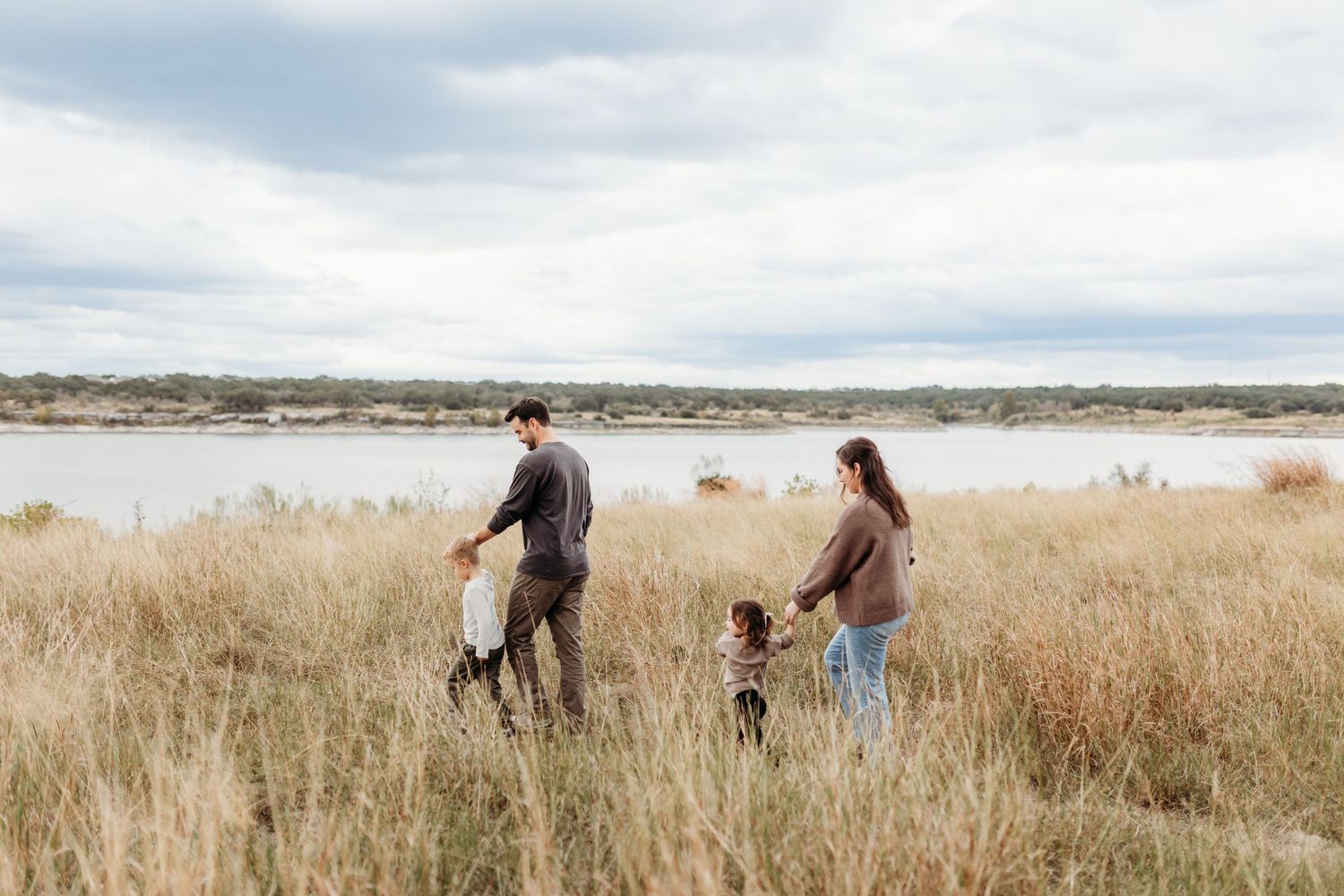 Family walking through tall golden grass near a serene lake on a cloudy day.