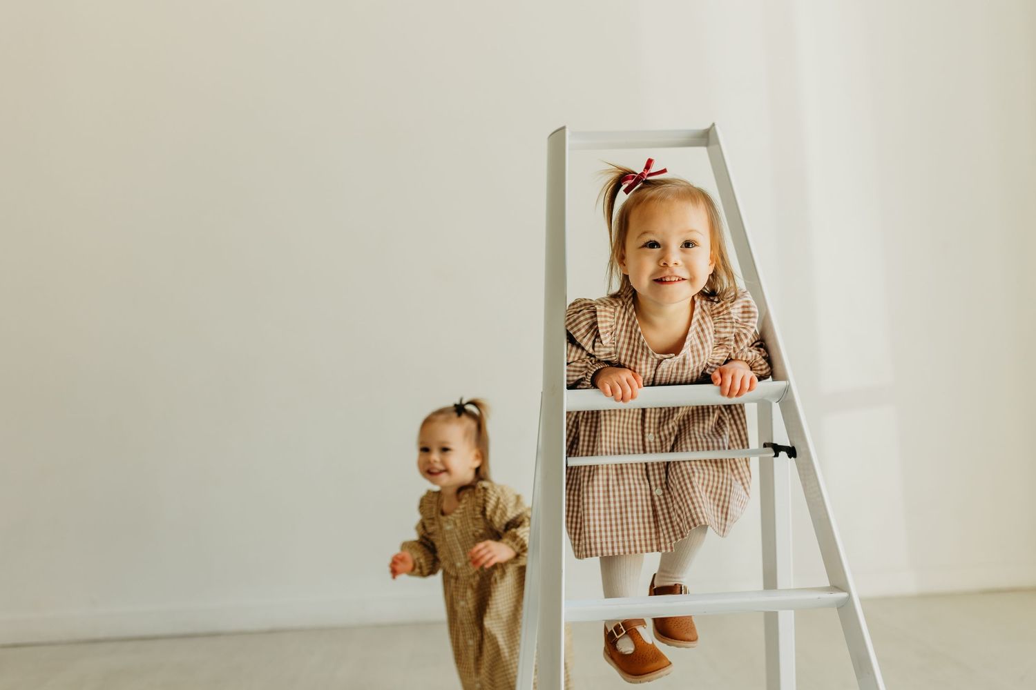 Two young sisters play on white ladder in bright minimalist studio during family photo session.