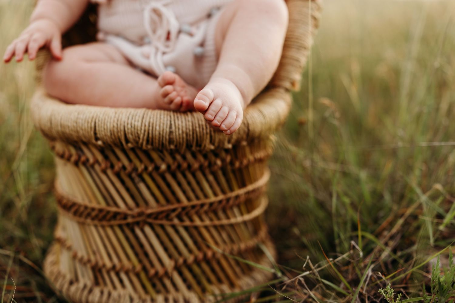 Baby sitting in wicker basket outdoors in grassy field wearing neutral romper.