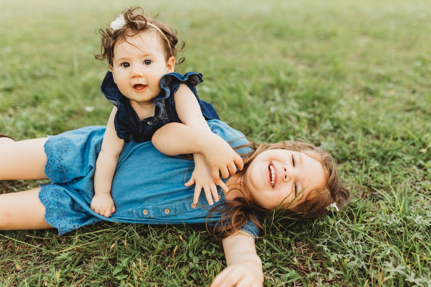 Two little girls in blue denim outfits playing together on lush green grass outdoors.