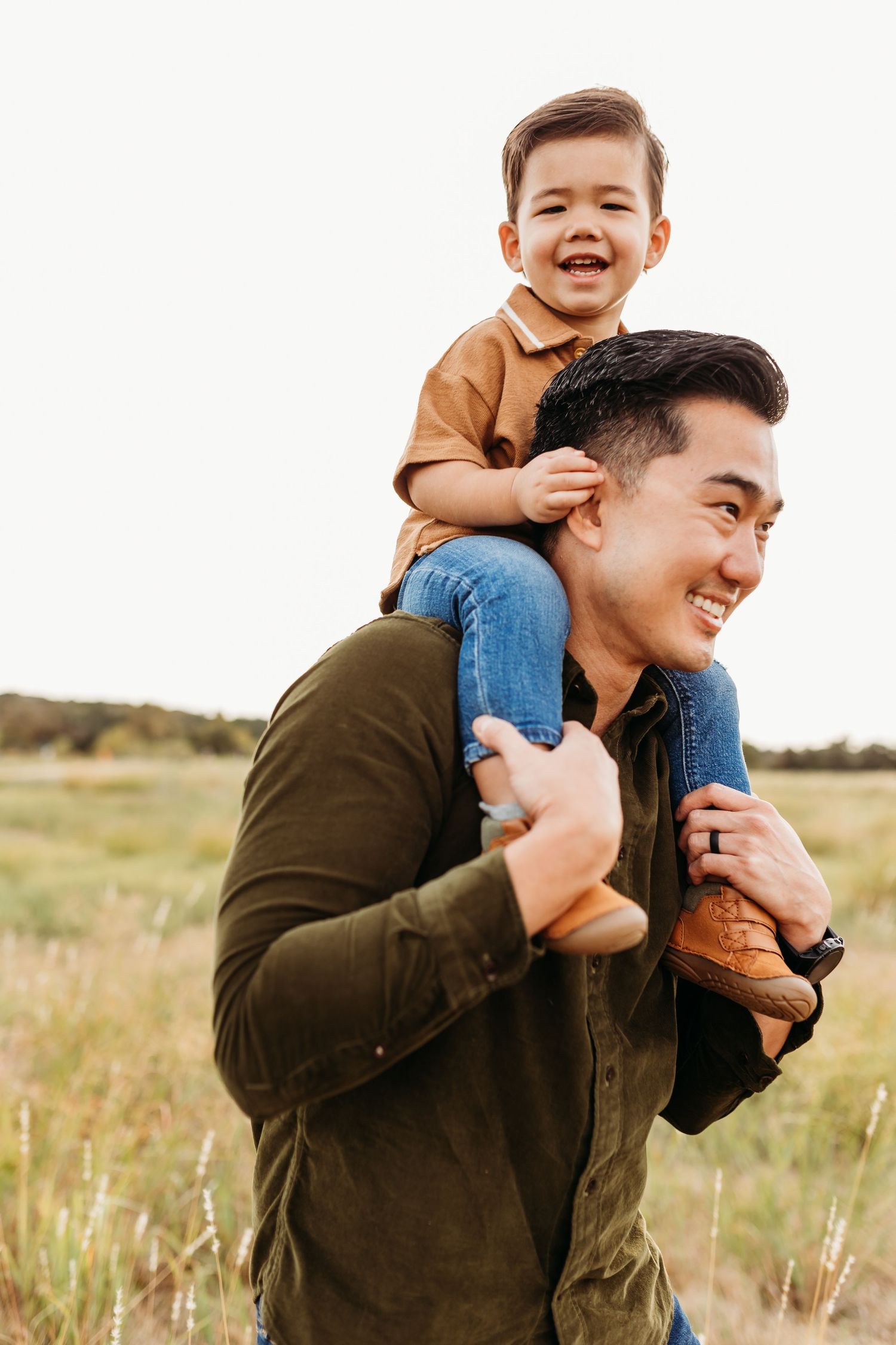 A smiling Asian father carries his laughing toddler son on his shoulders in an open grassy field.