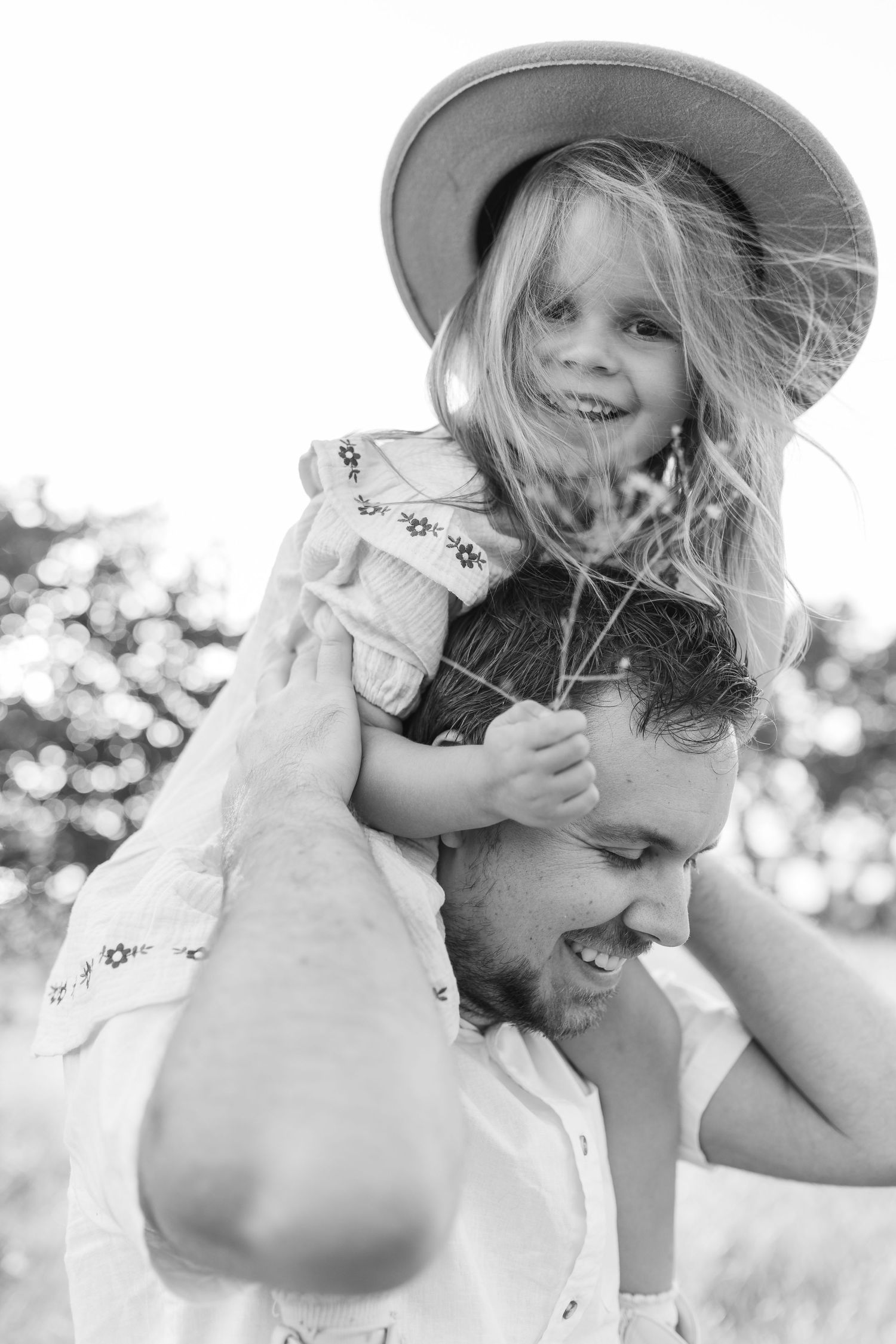 Black and white photo of smiling girl in hat riding on fathers shoulders outdoors.