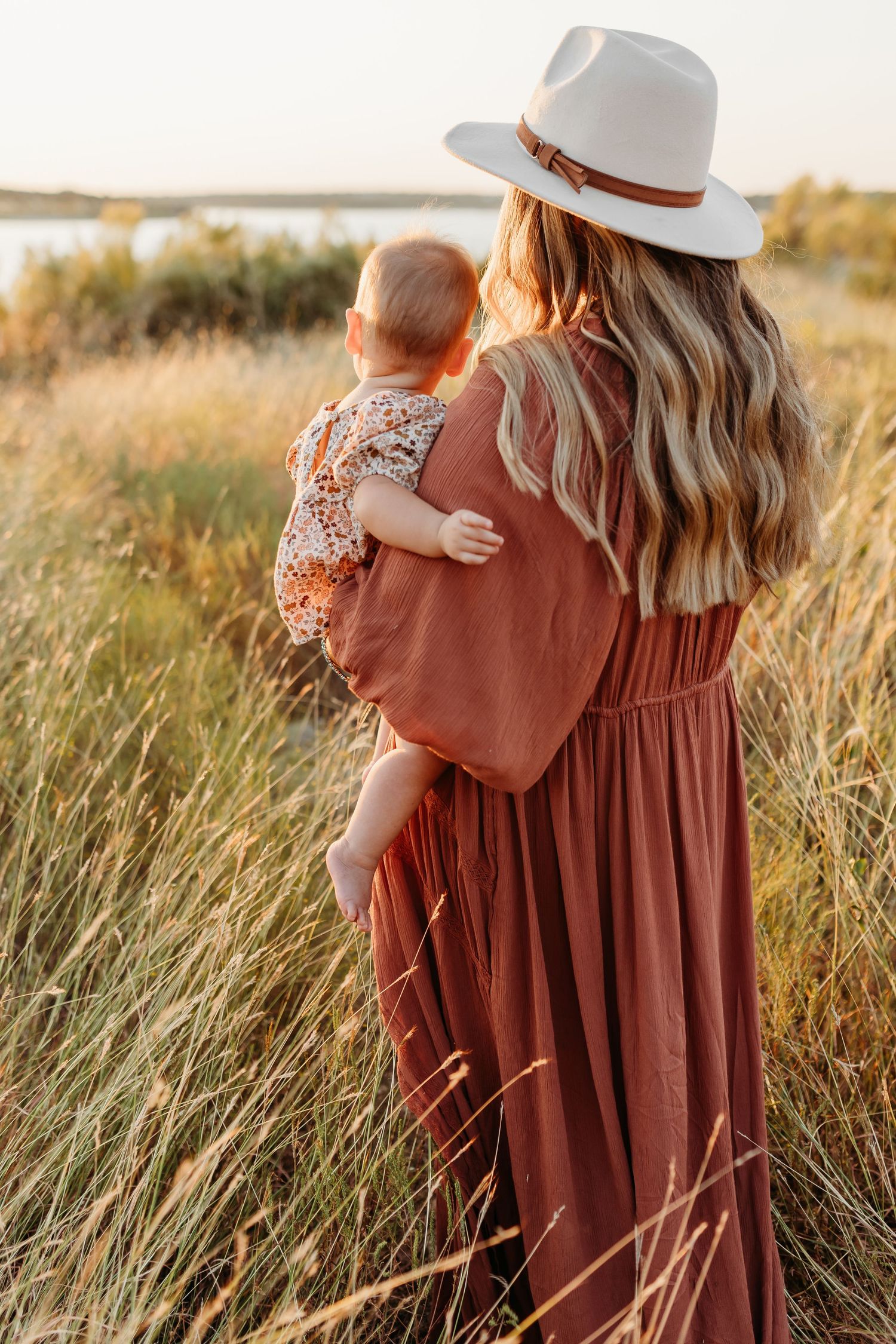 Mother in rust maxi dress and white hat holding baby while standing in tall golden grass at sunset.