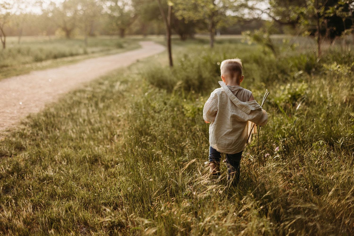 A young boy walks along a grassy country path surrounded by tall green grass at sunset.