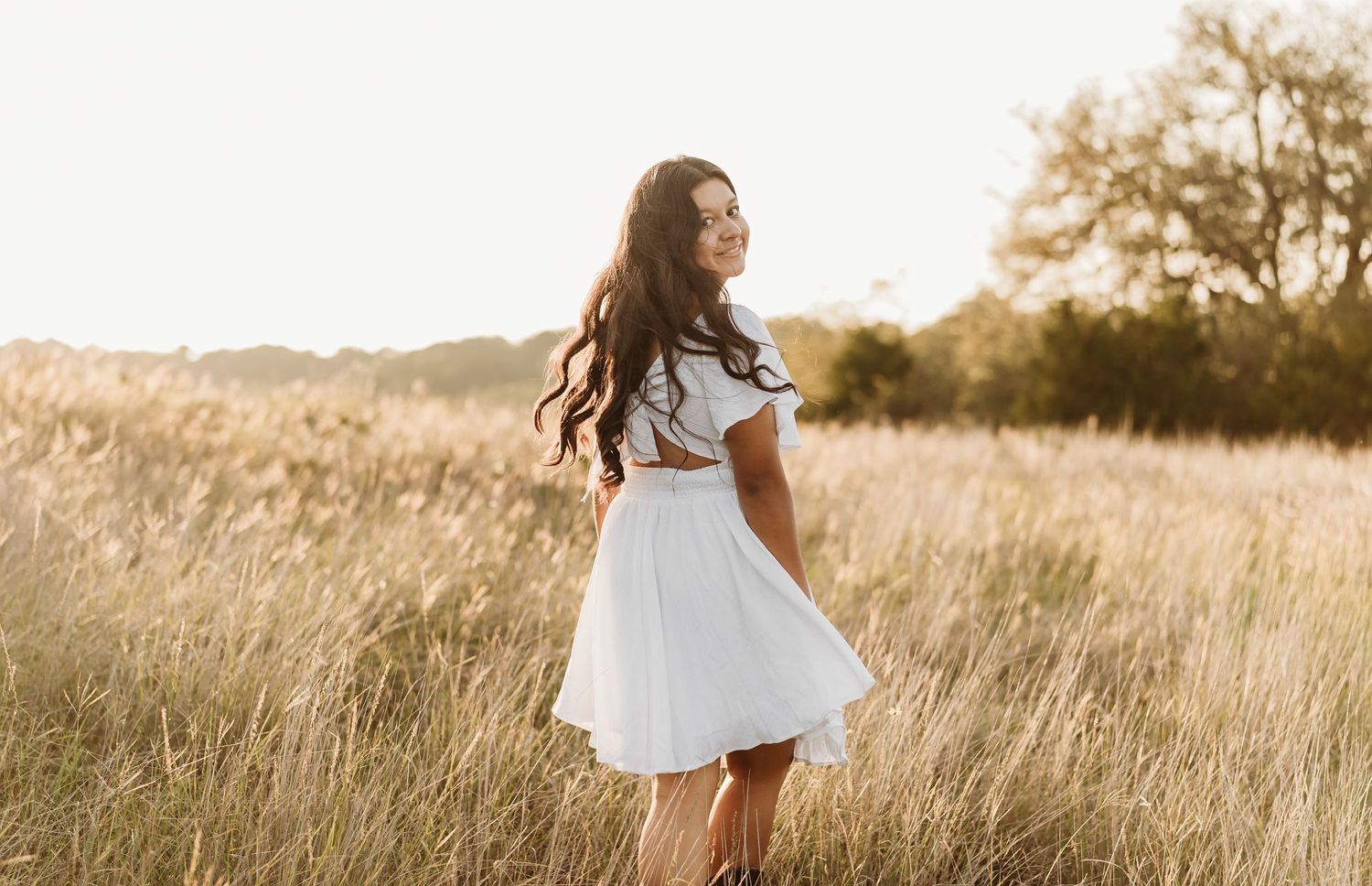 Young brunette woman in a white dress standing in a golden sunlit field during sunset portrait session.