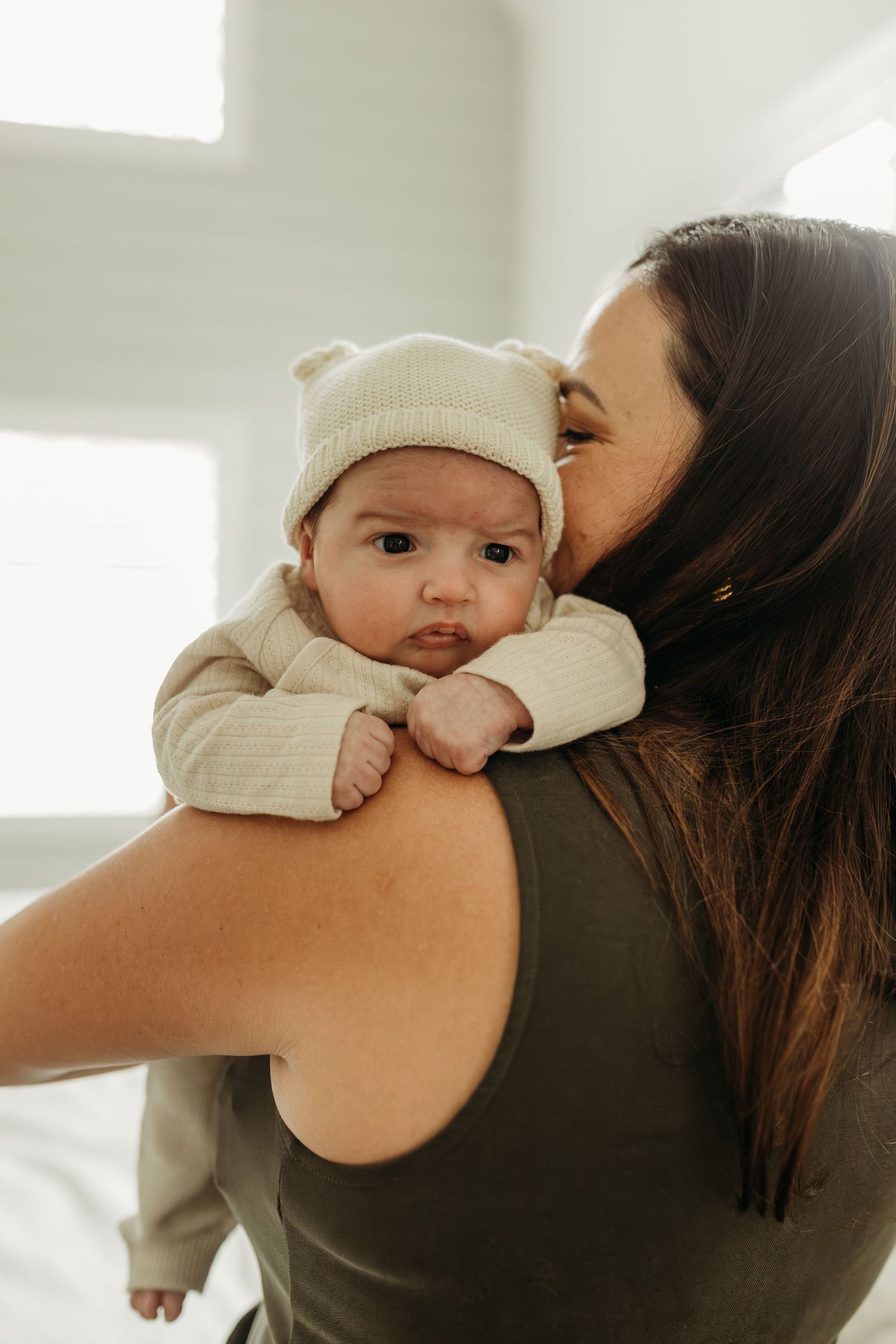 Mother in olive tank top kisses newborn baby wearing cream knit bear hat on her shoulder.