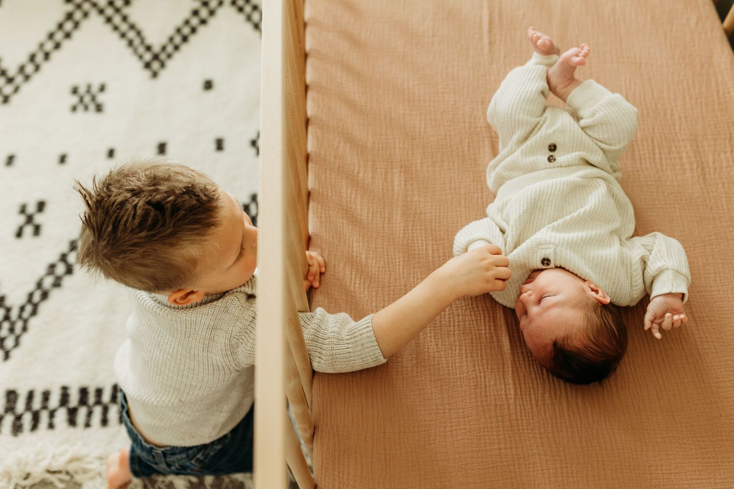 Toddler reaches into crib to touch newborn sibling lying on caramel crib sheet.