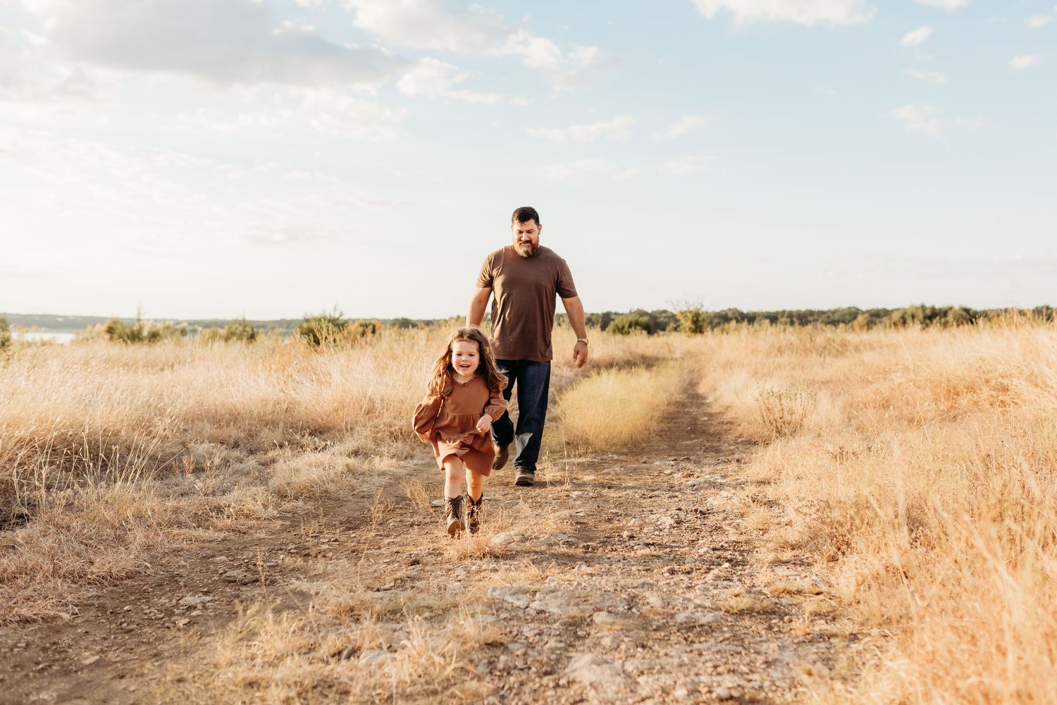 Father and young daughter running together on a dirt path through a golden dry grass field.