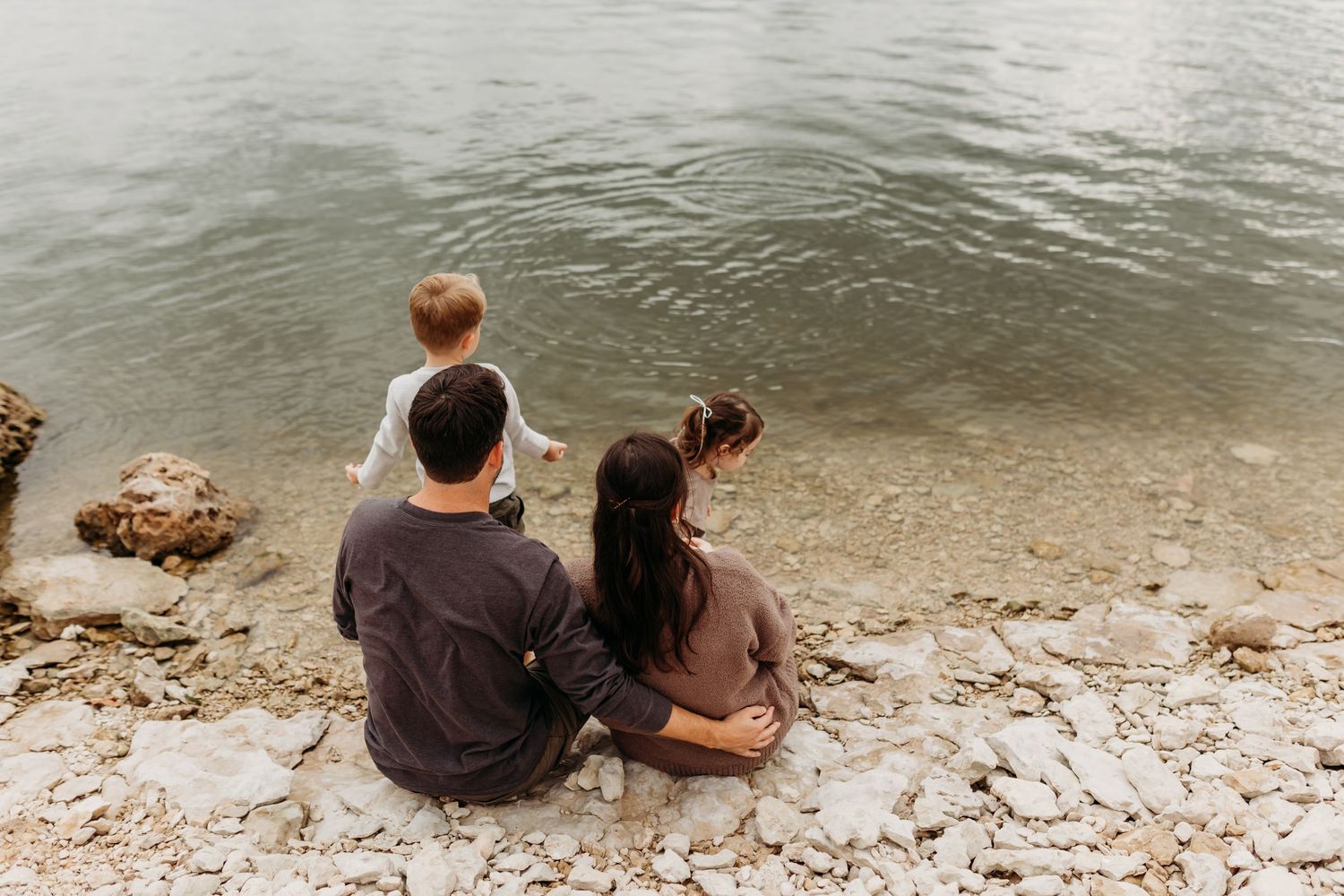Family sitting by rocky waterside, two children and parents enjoying peaceful lake view.