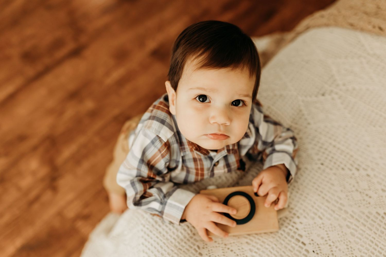Cute baby boy in plaid shirt lying on blanket looking up at camera indoors.