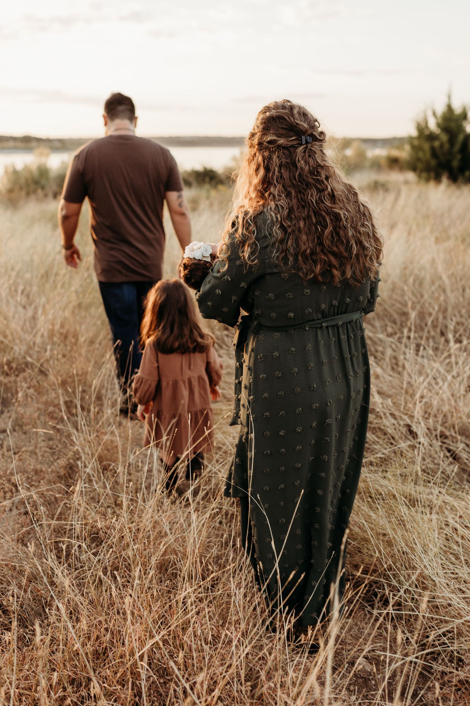 Family of three walking through dry golden grass field toward a lake at sunset from behind.