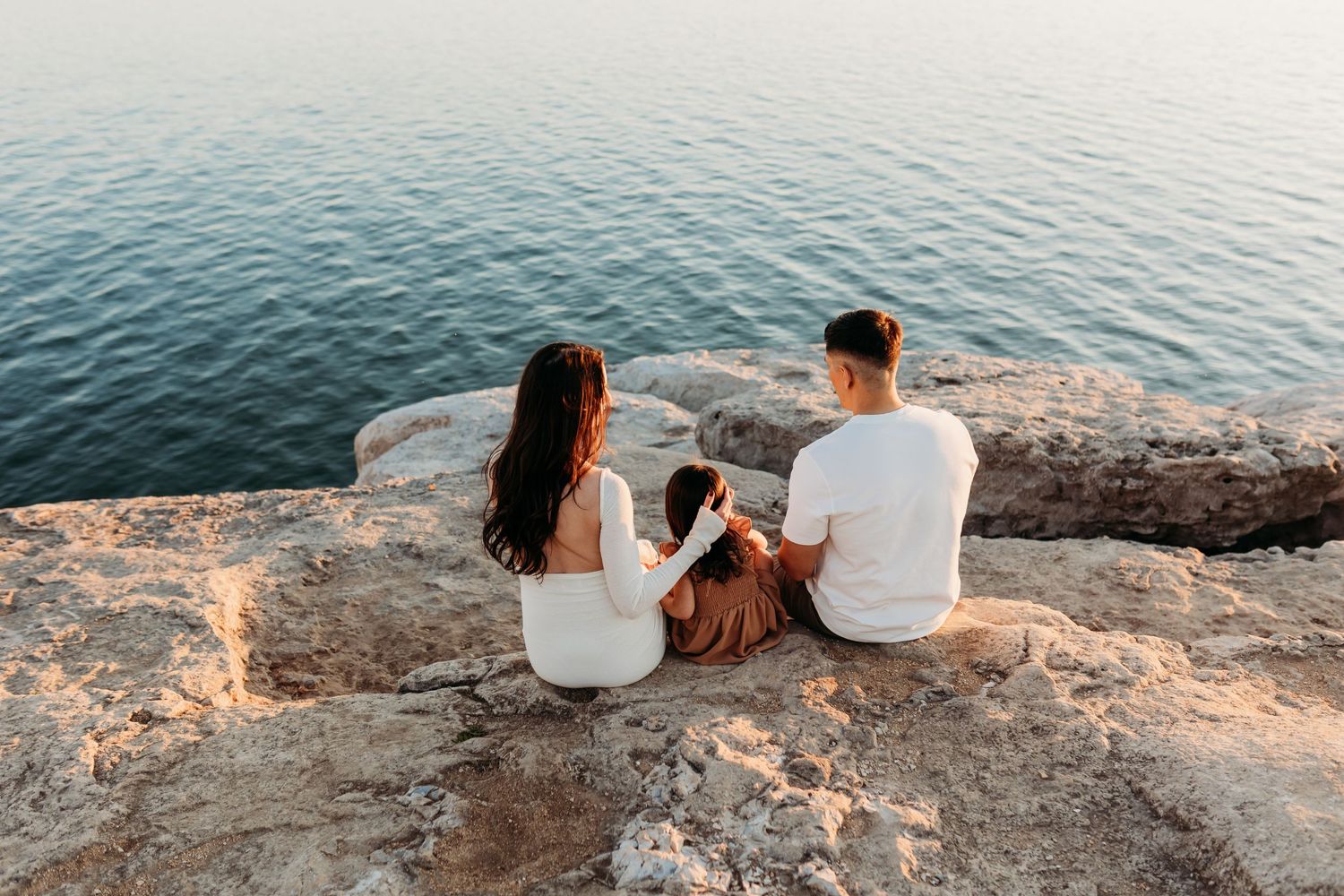 A family of three sits together on rocky cliffs overlooking calm turquoise water at sunset.