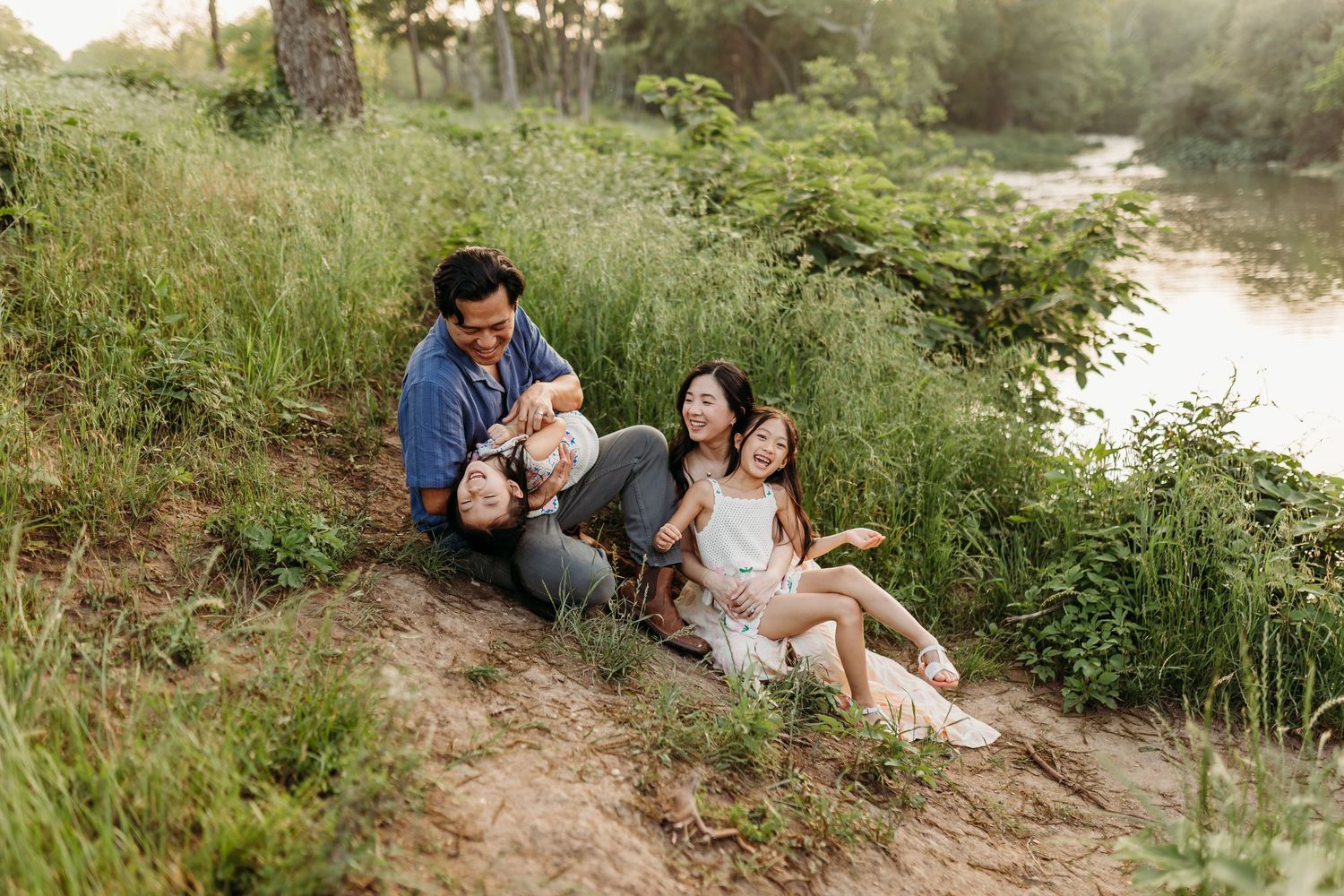 Joyful family sitting on riverbank surrounded by tall grass laughing together in warm sunlight.