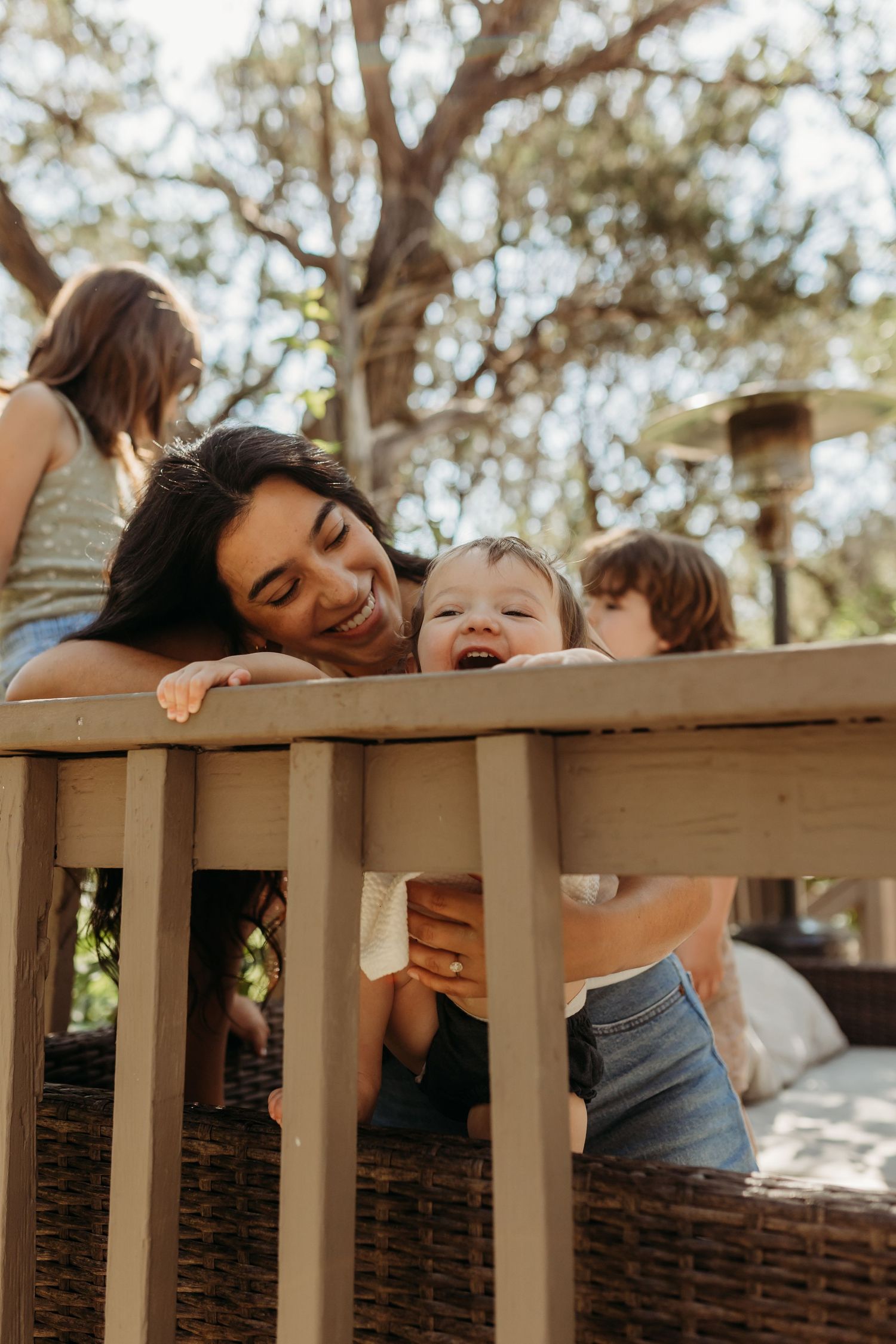 A mother smiles lovingly at her laughing toddler peeking through wooden deck railings outdoors.