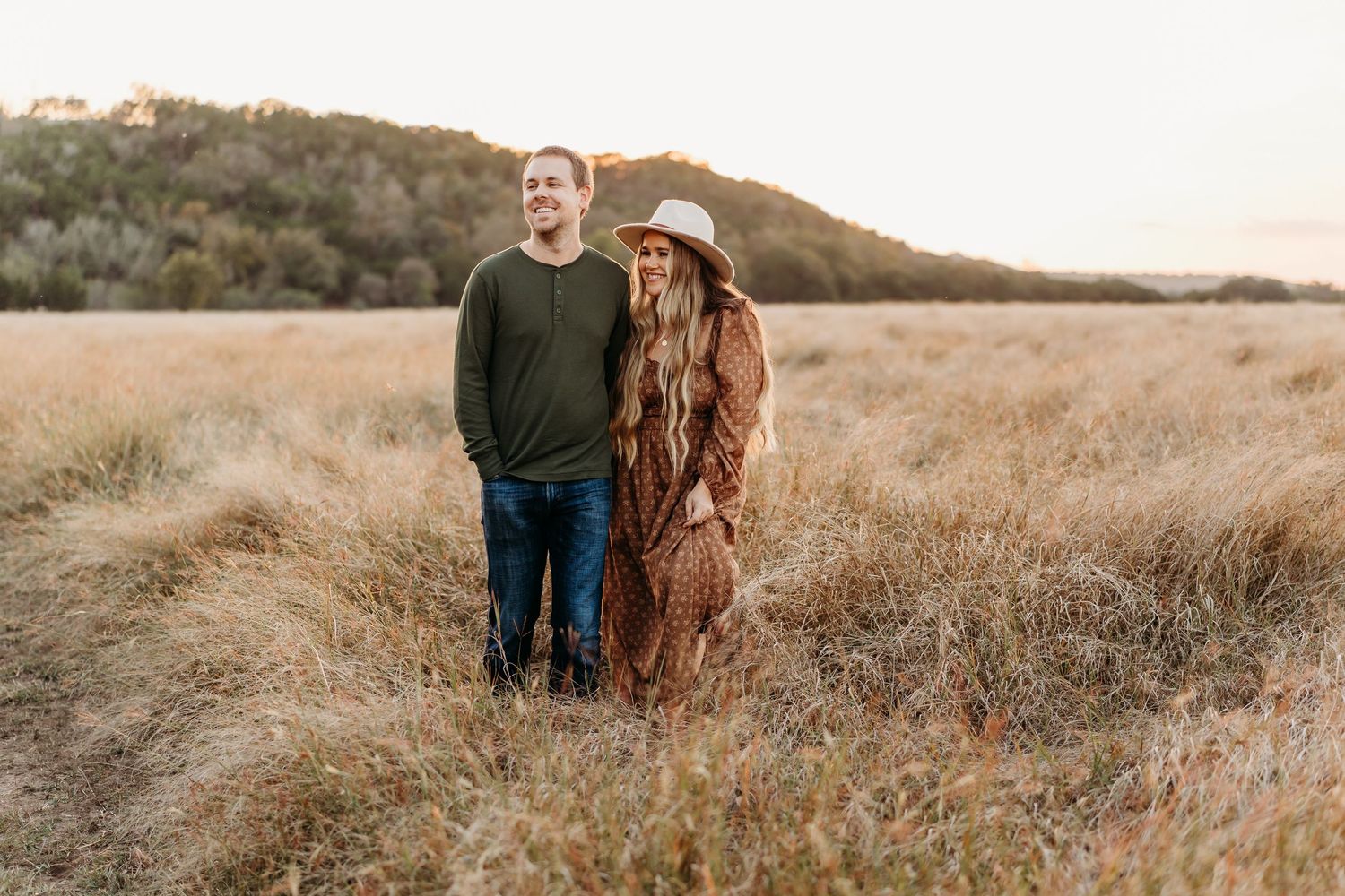 Couple standing together in golden dry grass field at sunset with hills in background.