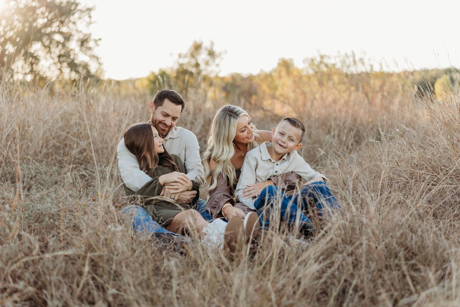Family of four sitting and laughing together in a golden grassy field at sunset.
