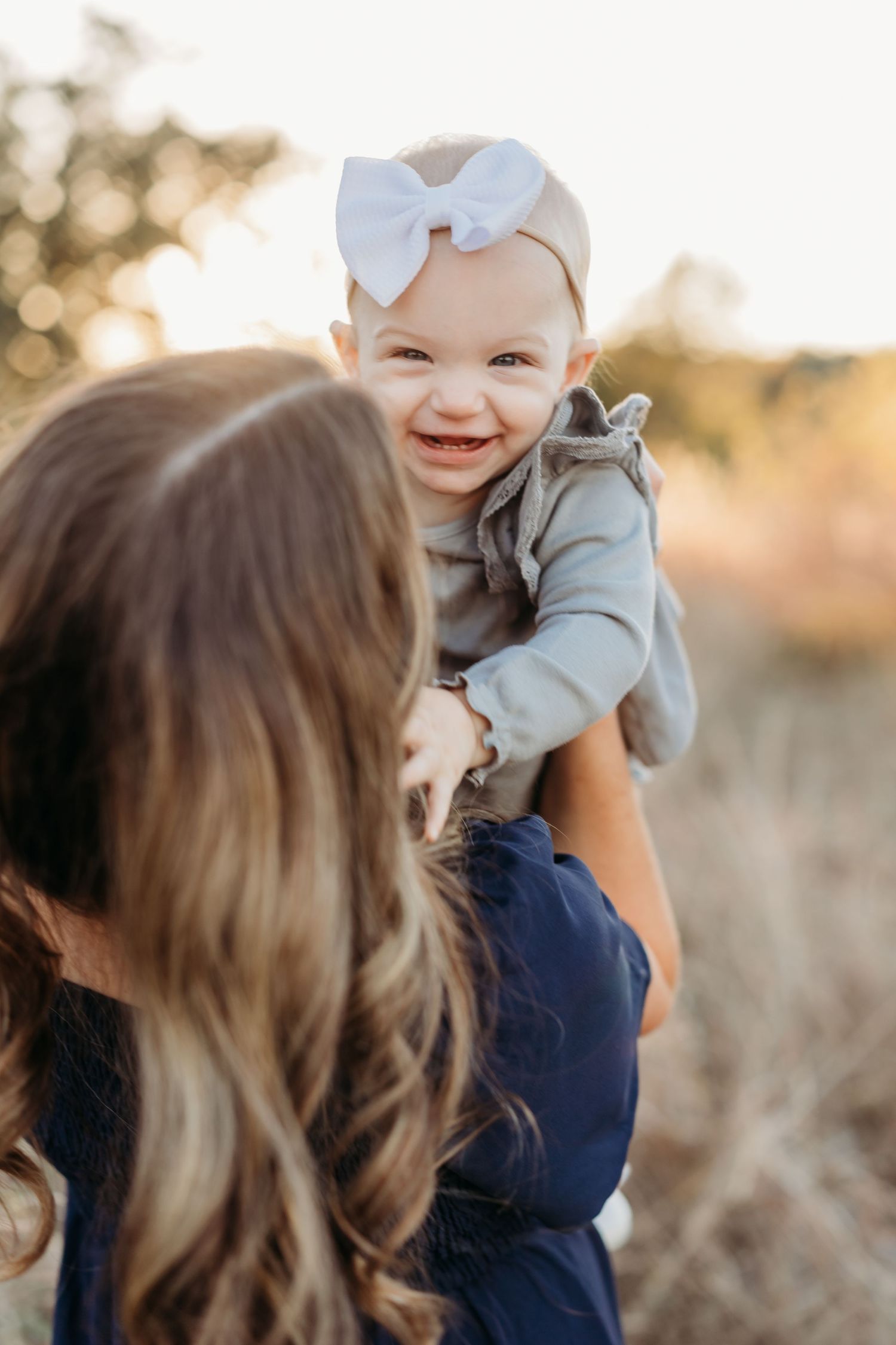 Smiling baby girl with a white bow headband being held up by her mother outdoors.