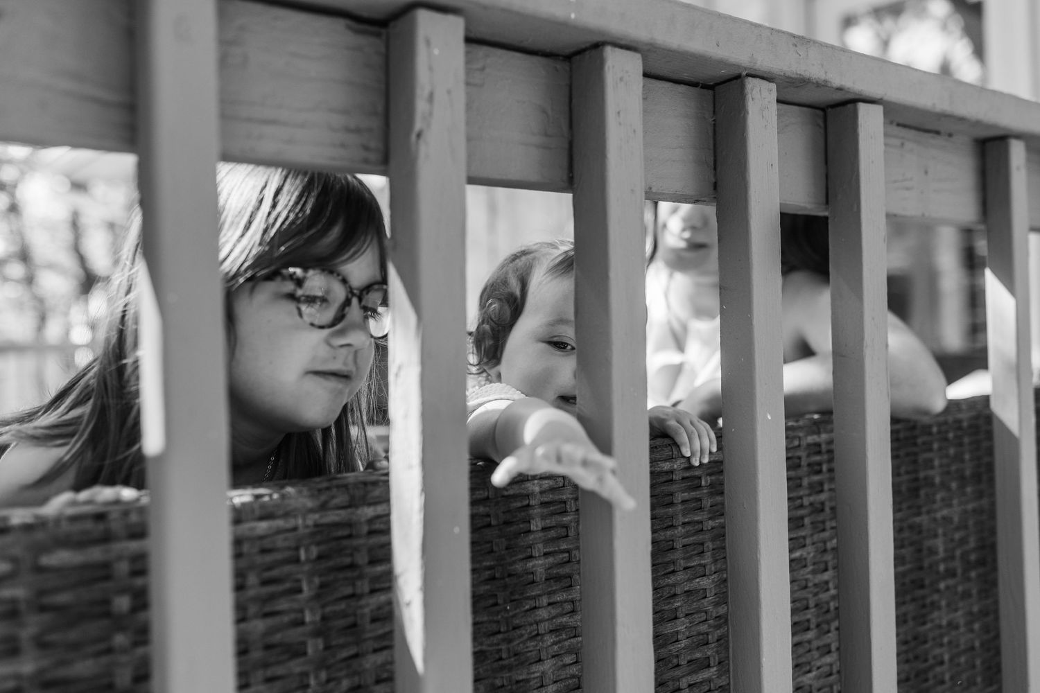A black and white photo of a girl with glasses and a baby peeking through wooden deck railings.
