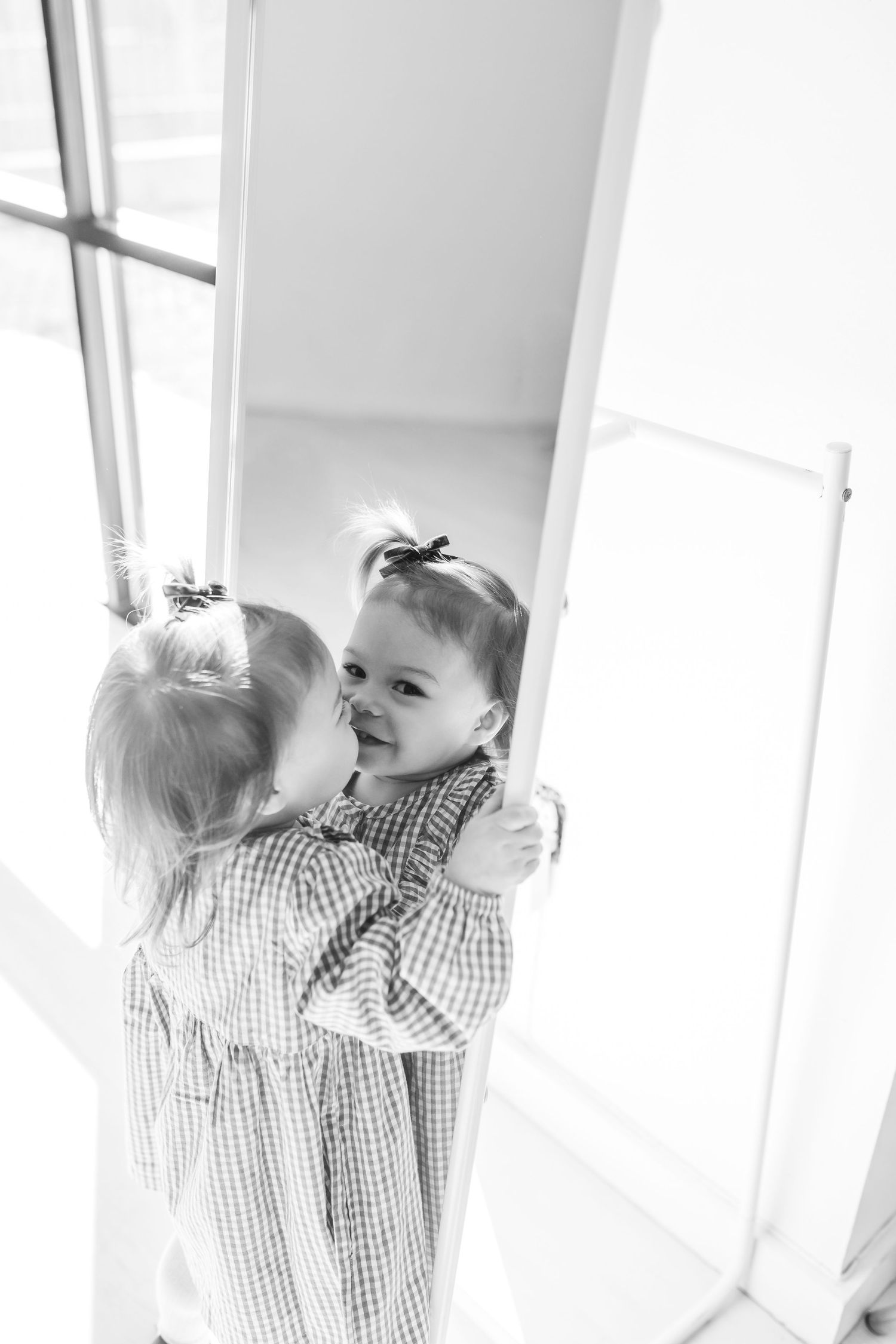 Young toddler girl in plaid dress looks at her reflection in a mirror in black and white photo.