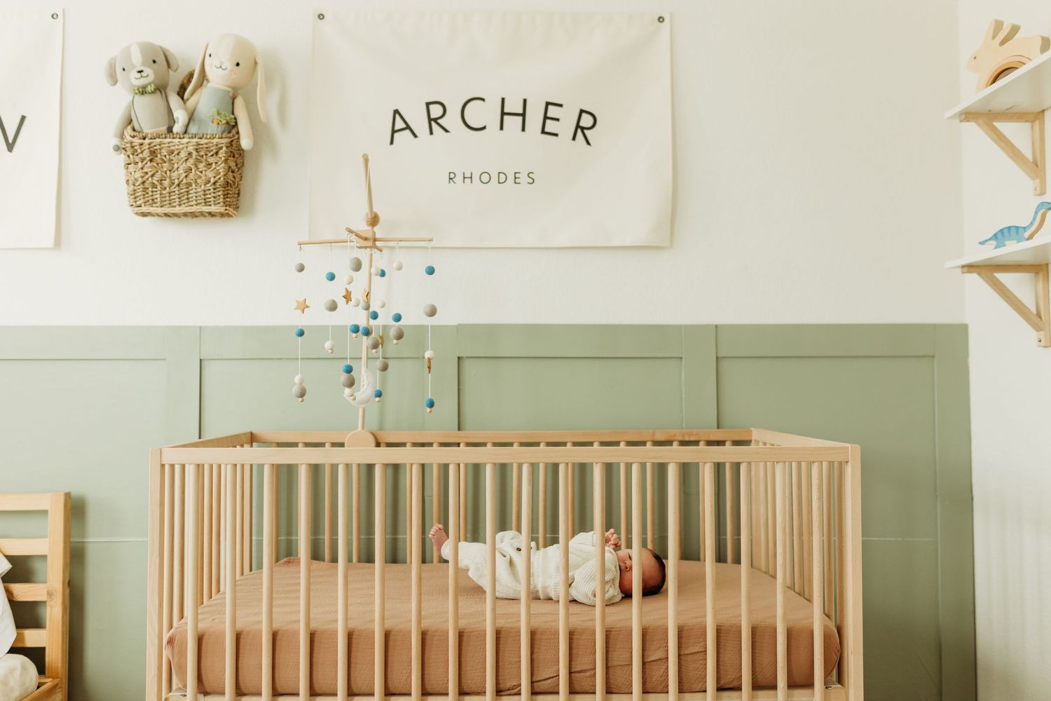 Newborn baby sleeps in wooden crib beneath Archer Rhodes name banner in sage nursery.