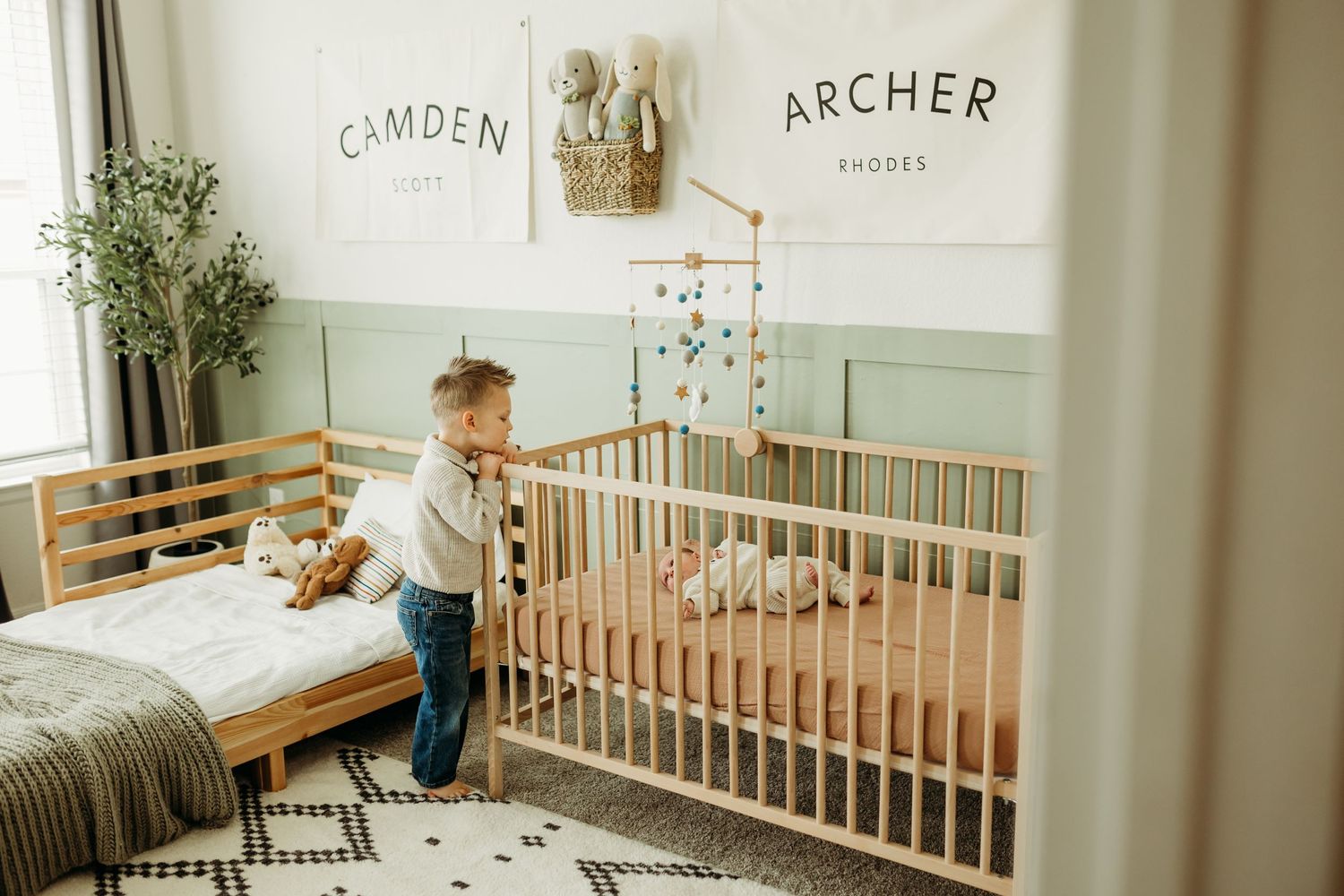 Toddler stands in shared nursery between Camden and Archer name signs above beds.
