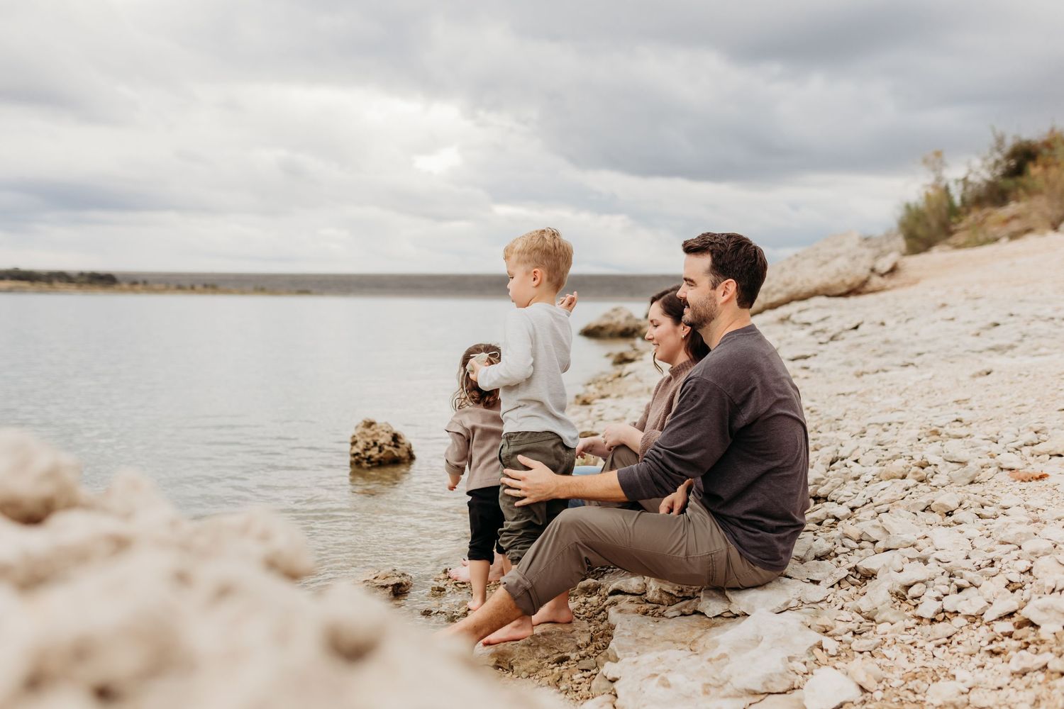 Family sitting together on rocky shoreline watching children play near calm lake water.