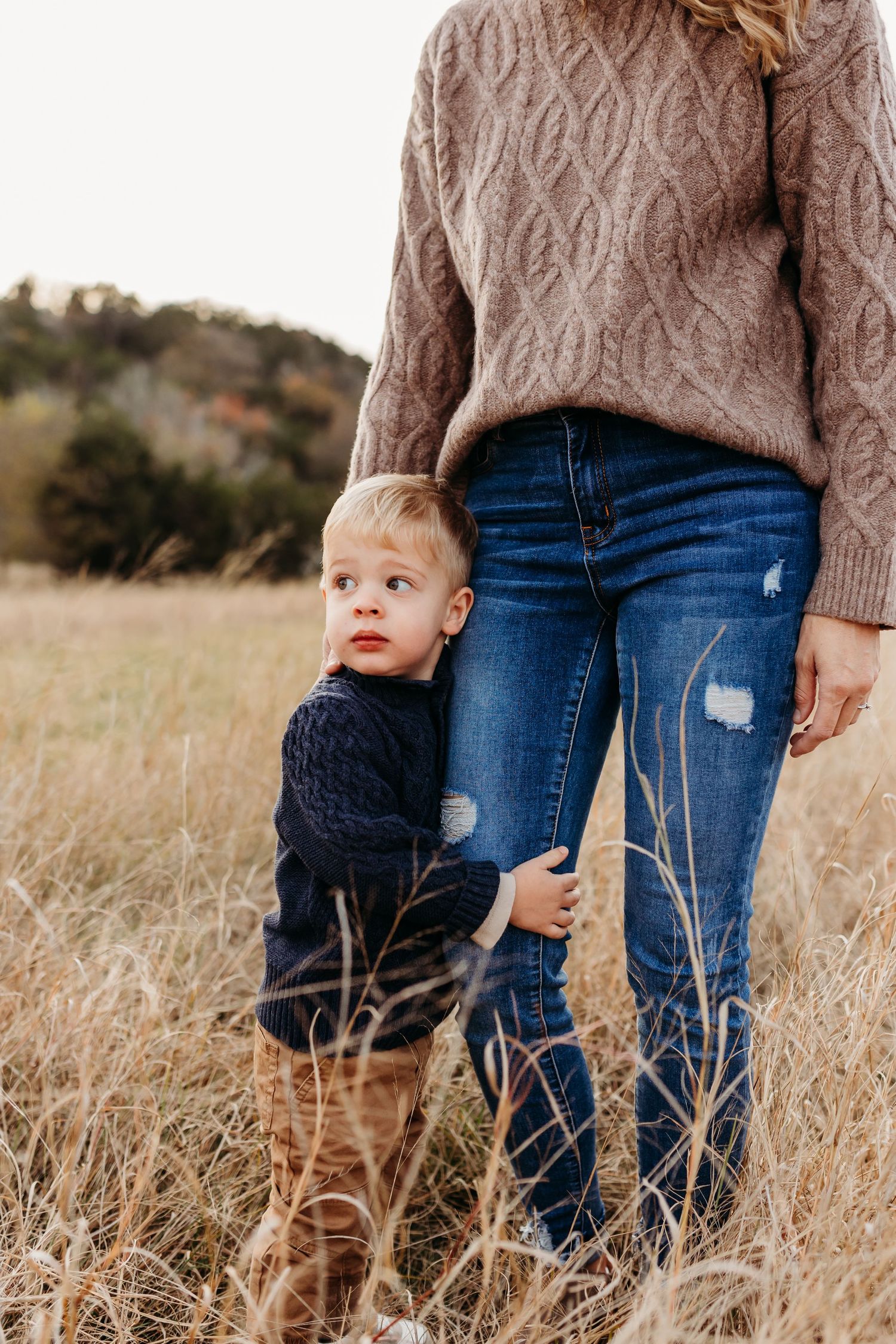 A young toddler boy clings to his mother's leg in a golden field during an outdoor fall family photo session.
