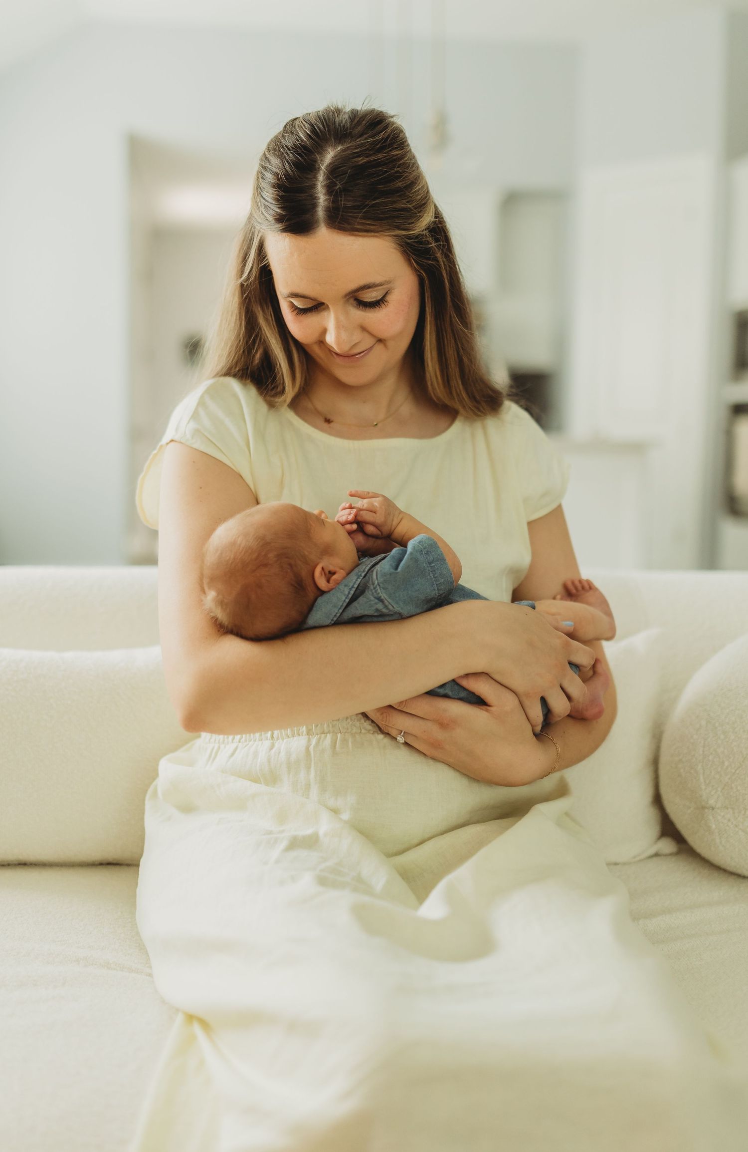 Young mother in white dress tenderly holding sleeping newborn baby on white sofa.
