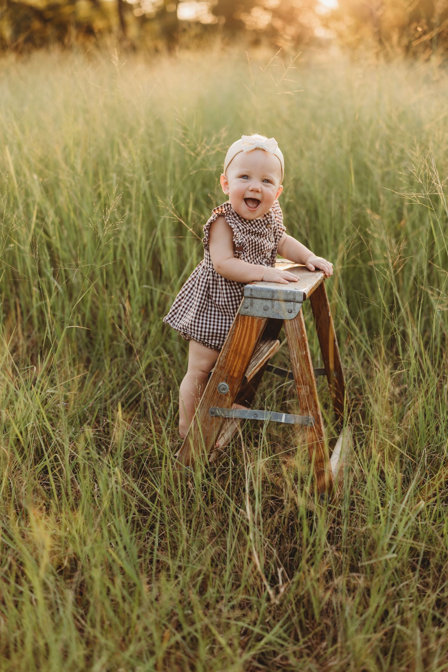 Joyful baby girl in checkered dress laughing while standing on wooden ladder in field.