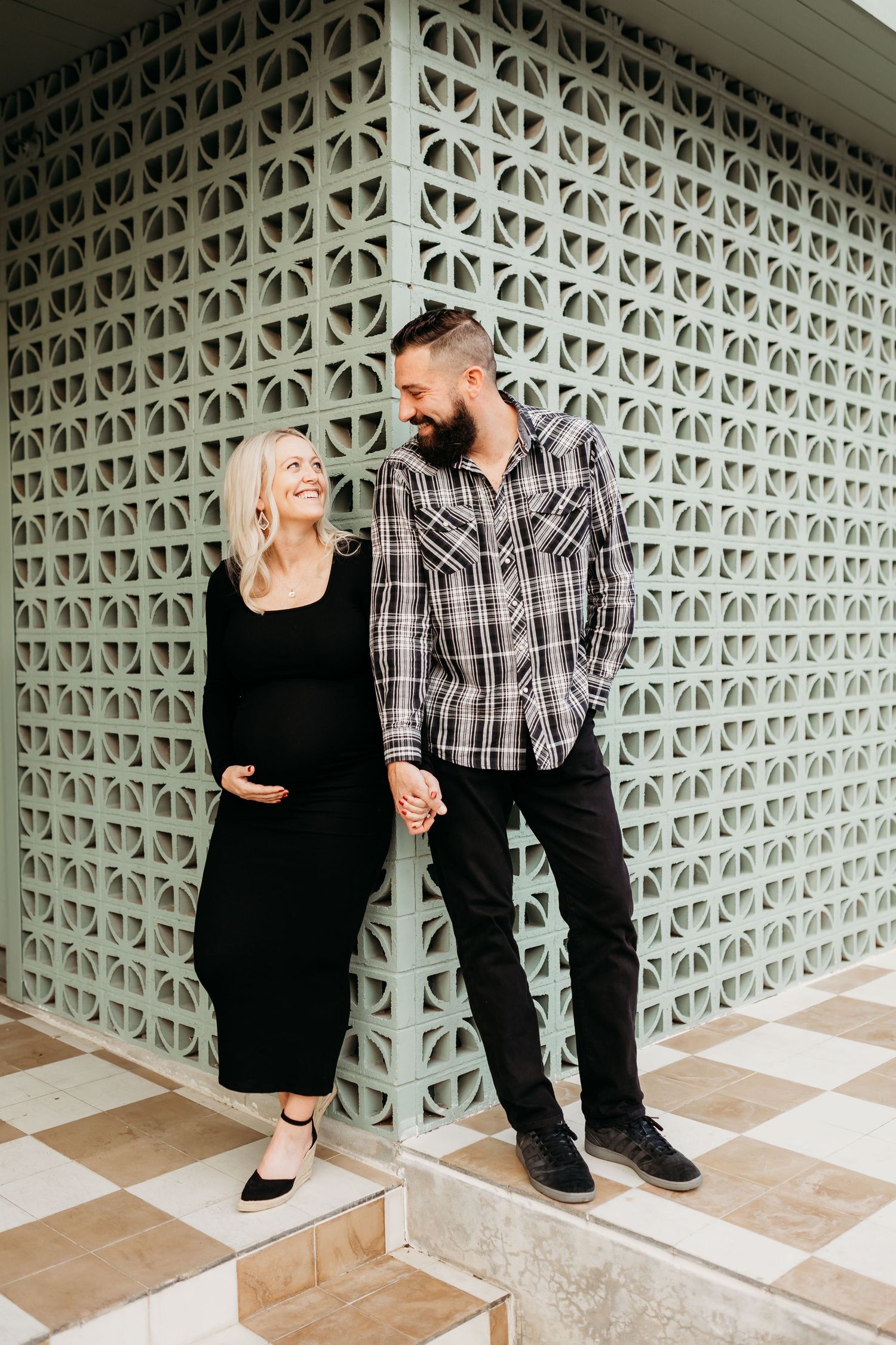 A pregnant woman in a black dress holds hands with her bearded partner near a decorative lattice wall.