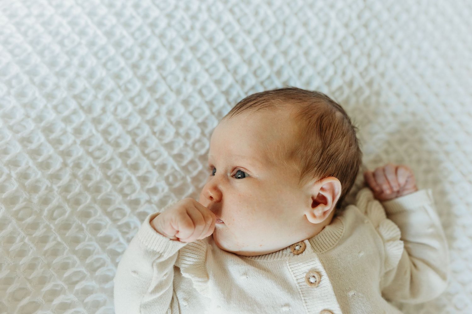 Close-up of a newborn baby in a cream outfit lying on a white textured blanket.