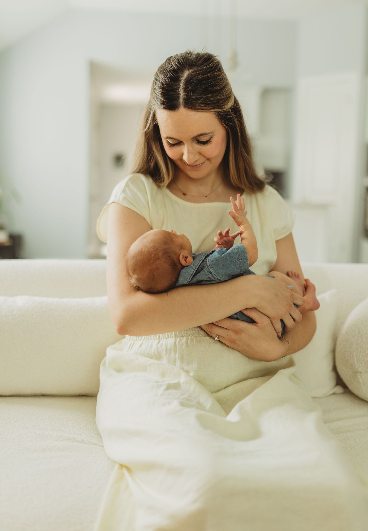 Young mother in cream dress gently cradling newborn baby while seated on white sofa indoors.