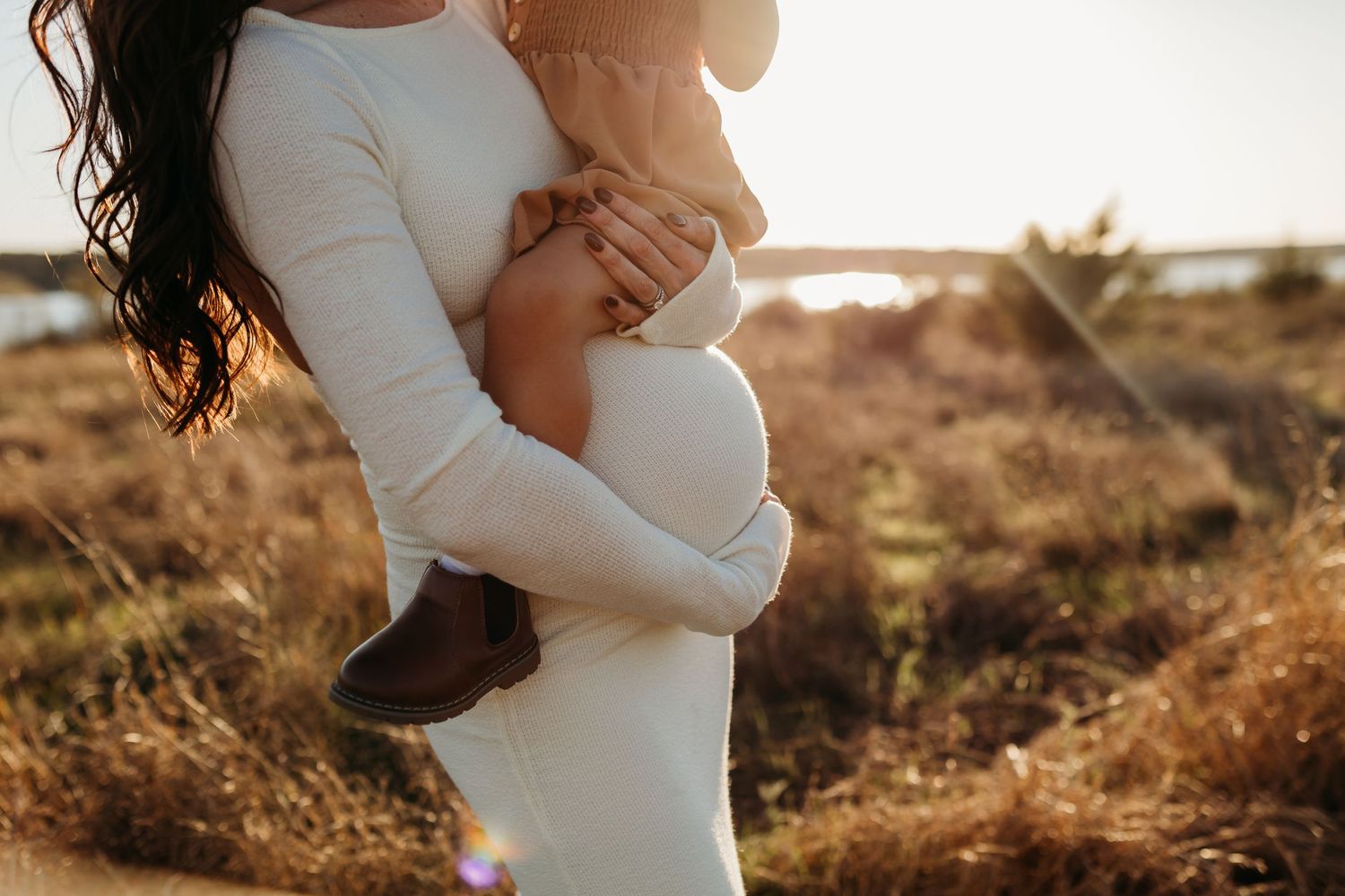 Pregnant woman in white dress holding a toddler close in a golden sunlit grassy field.