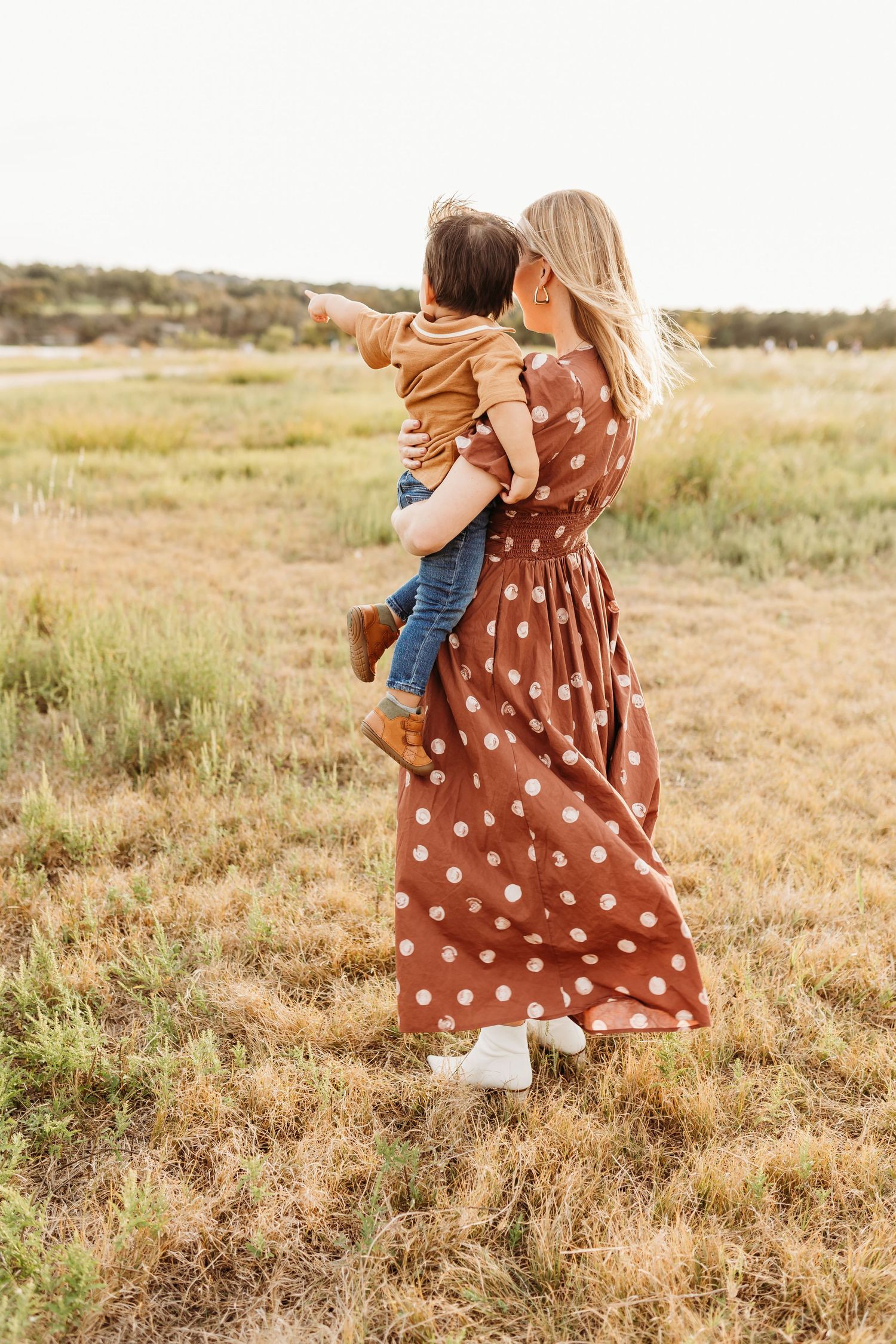A blonde mother in a brown polka dot dress holds her toddler son while he points into the distance in a field.