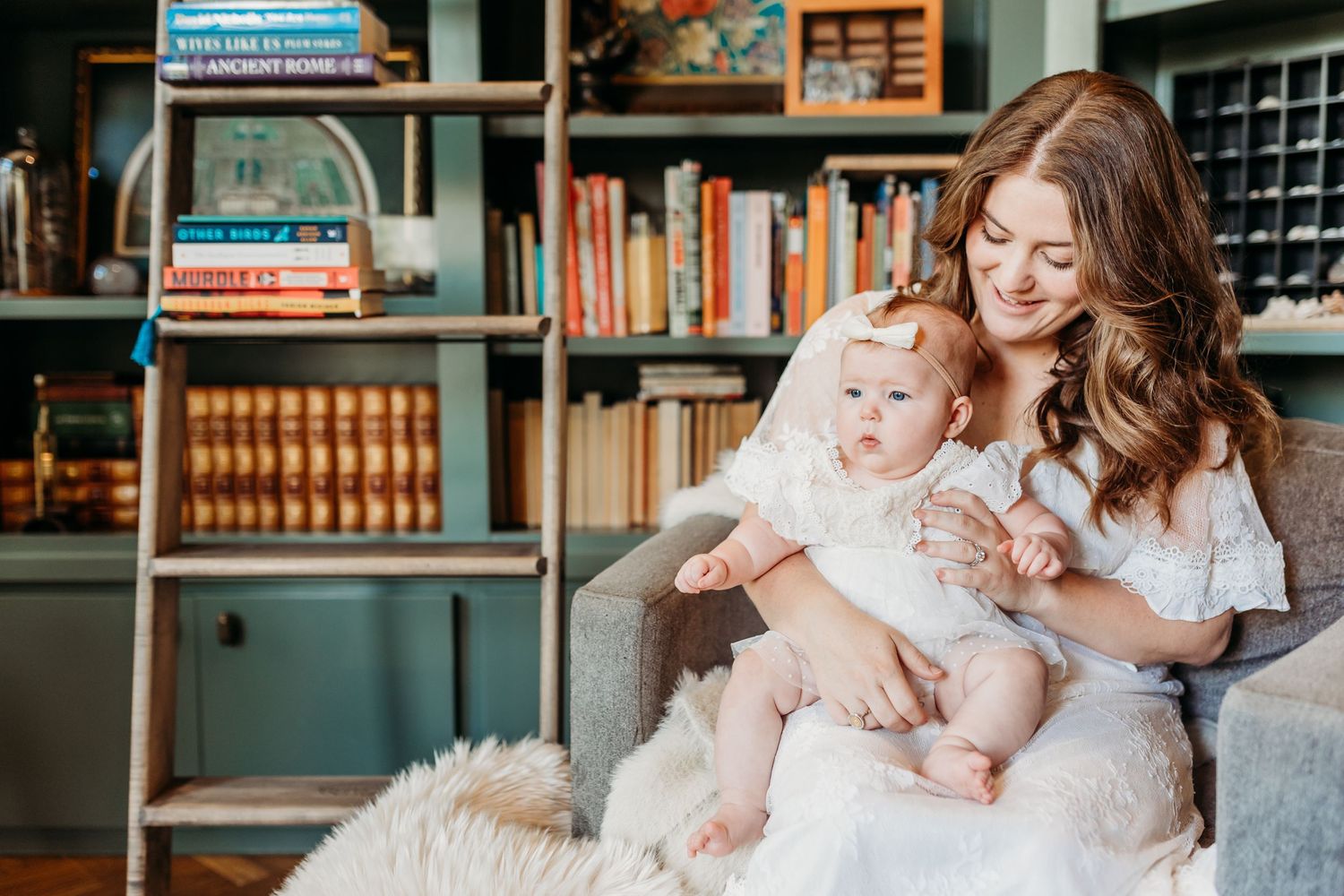 Young mother in white dress sitting in armchair holding baby girl near bookshelf.