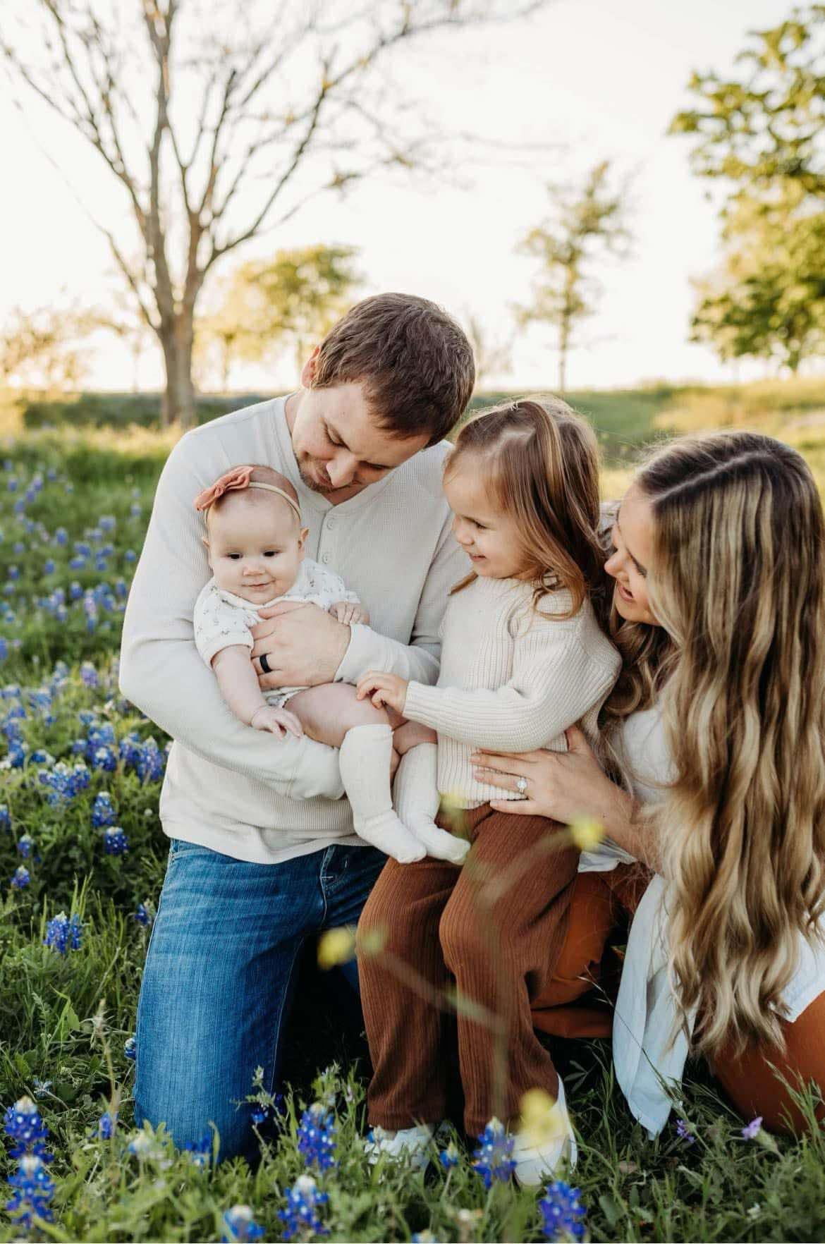 A happy family of four poses together in a blooming bluebonnet field at sunset.