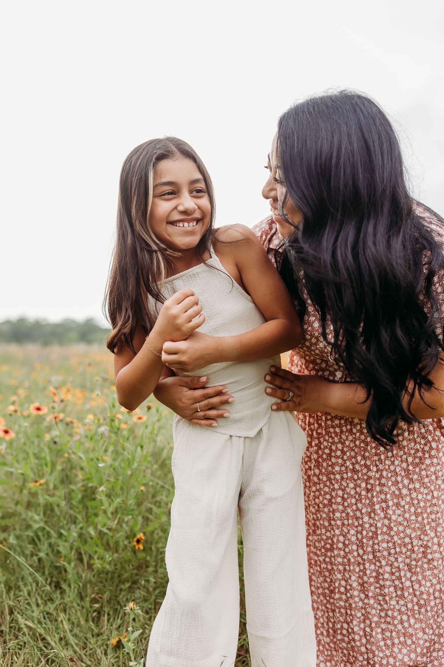 A mother hugs her laughing daughter in a wildflower field, both wearing neutral tones.
