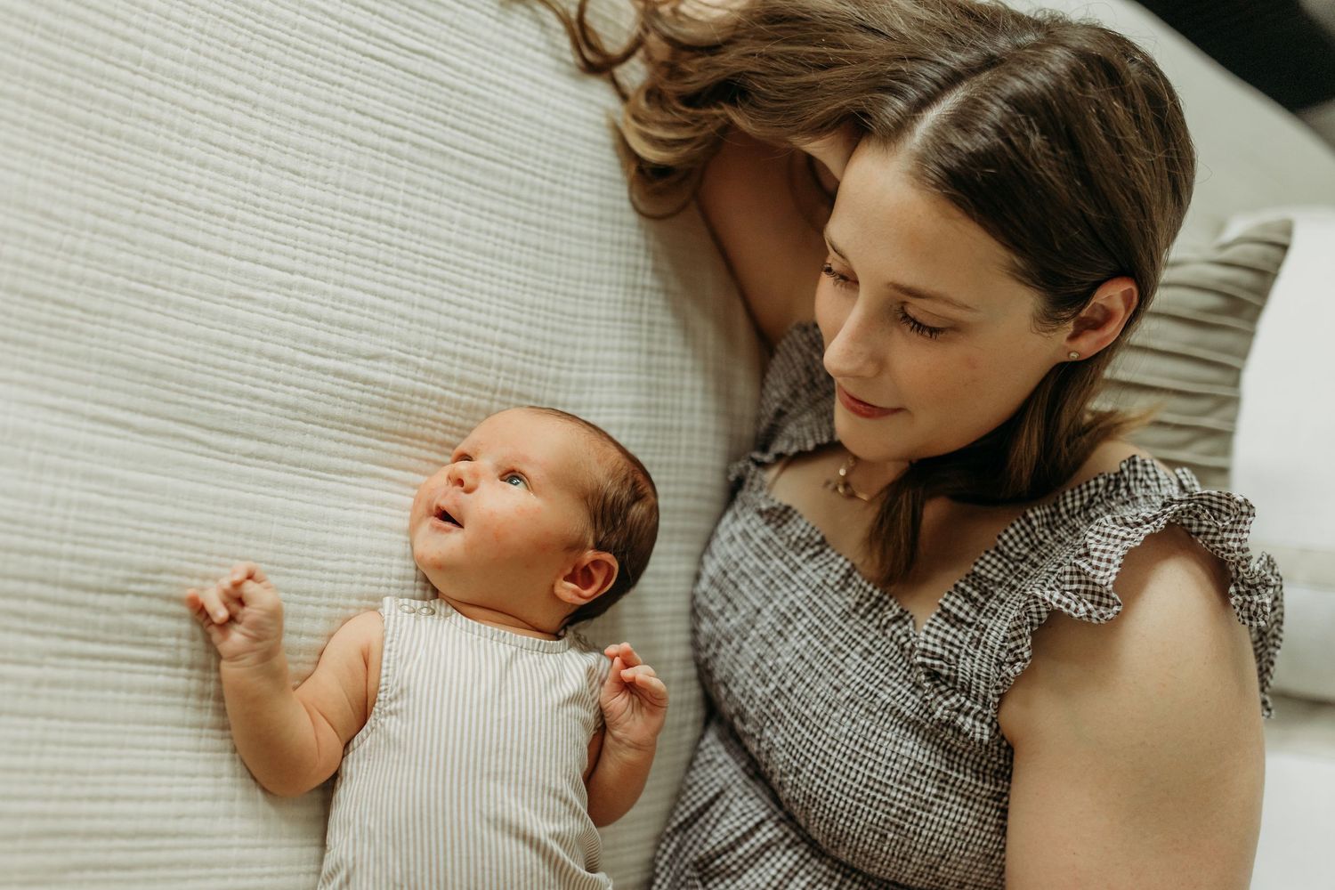 Overhead view of a mother lying beside her alert newborn baby on a white bed indoors.