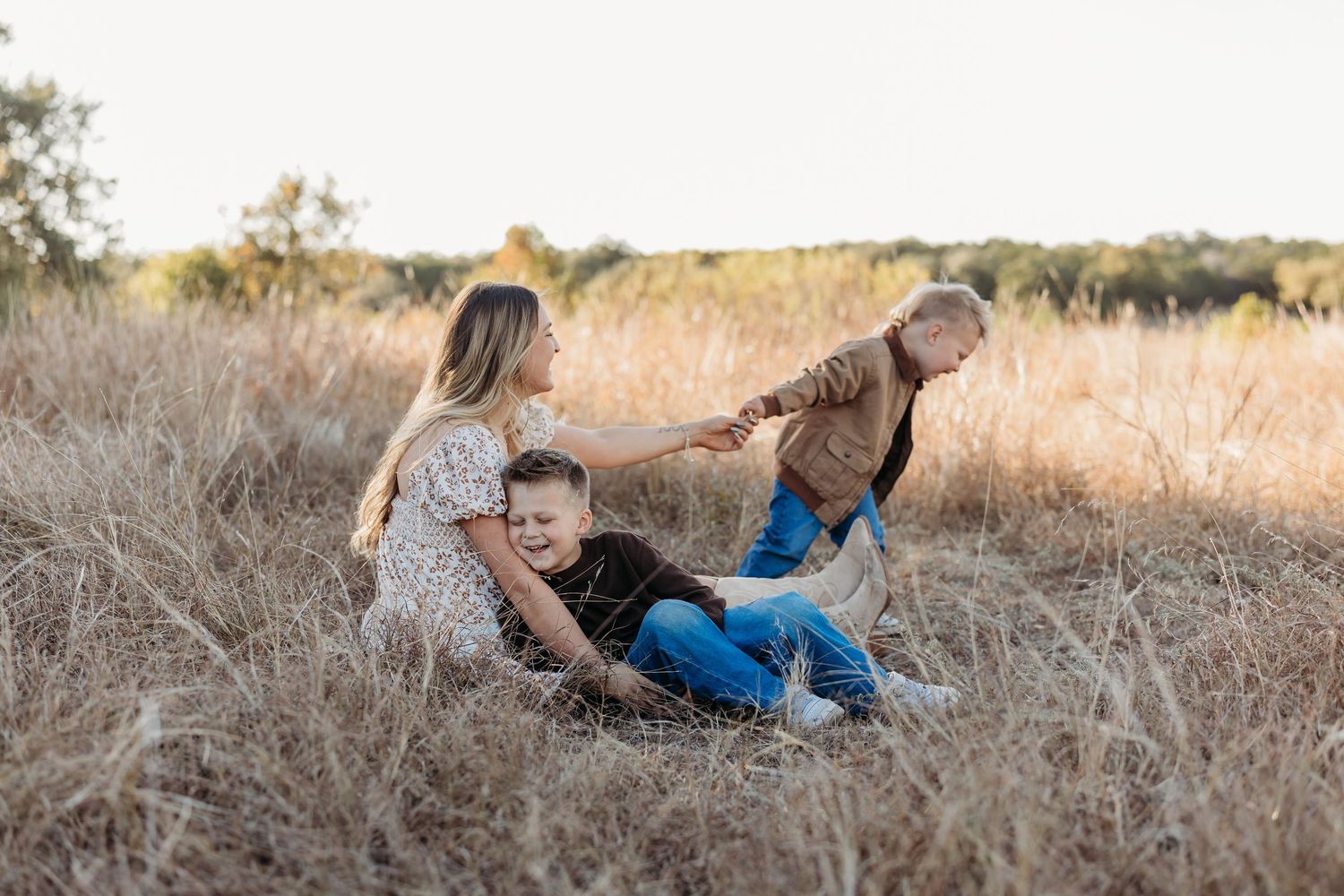 Mother sitting in a golden field with two young boys playing around her at sunset.