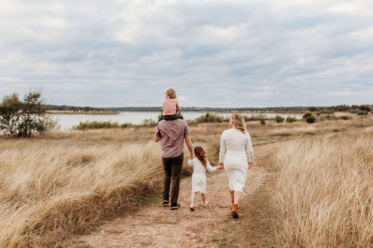 Family of four walking hand in hand on a trail through tall grass toward a lake.