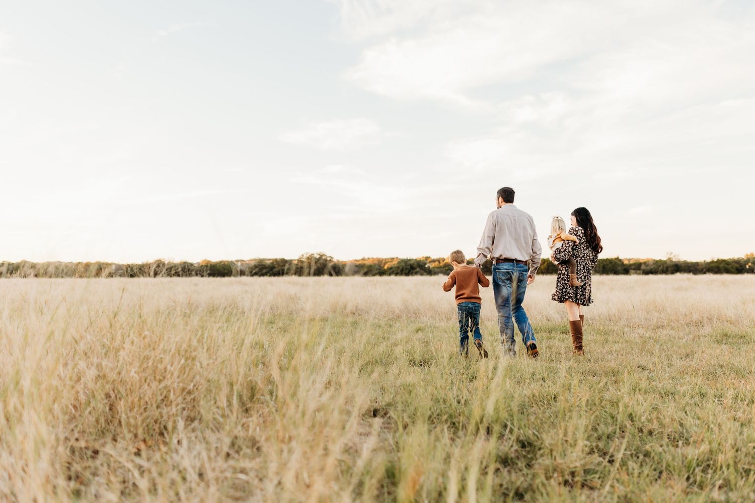 A family of four walks hand in hand through a wide open golden meadow under a bright sky.