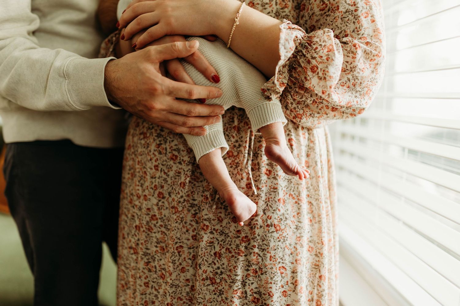Parents cradle their newborn baby near a window, mother wearing a floral dress.